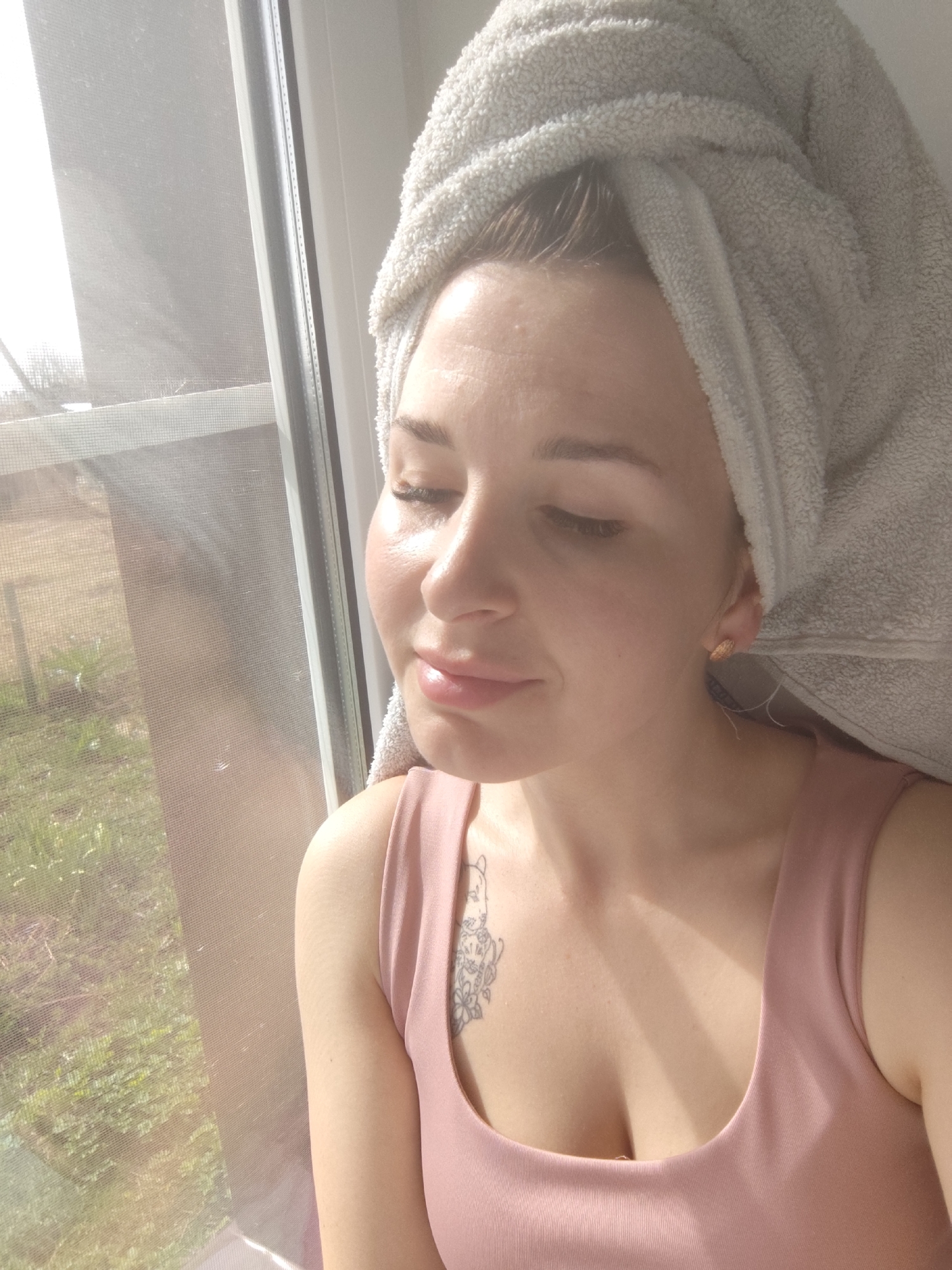 A woman with a towel wrapped around her head is seated near a window. She has fair skin, light-colored hair, and is wearing a pink top. A tattoo is visible on her upper chest. The window looks out onto a green, grassy area, and a building is visible in the distance. The location in the city of Фоки, Russia is not discernible from the image.