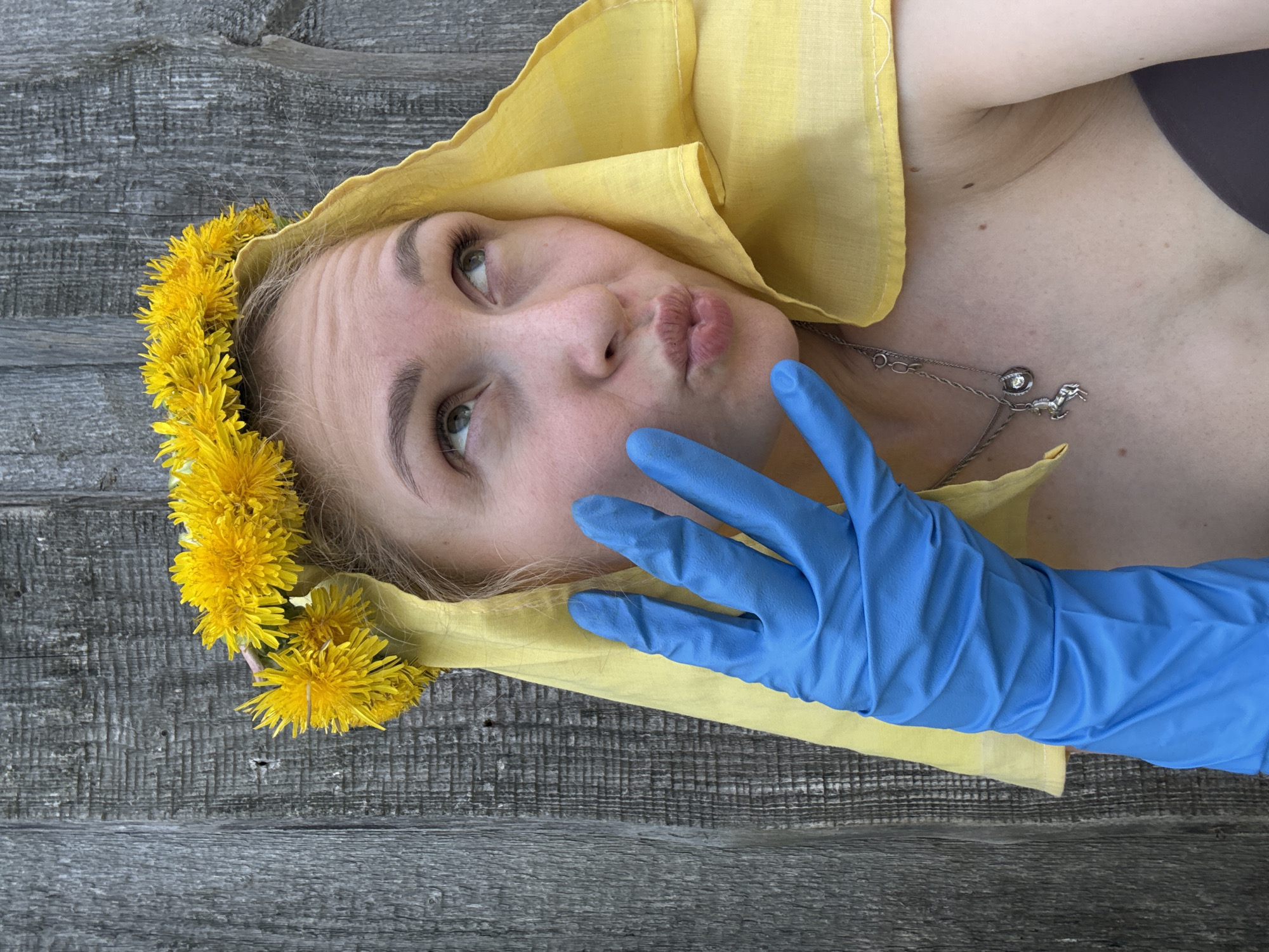 A woman lies on a gray, weathered wooden surface. She wears a dandelion wreath and a yellow cloth on her head, and a silver necklace. Her face is puckered into a kiss. A blue-gloved hand is near her face. No city location is indicated in the image.