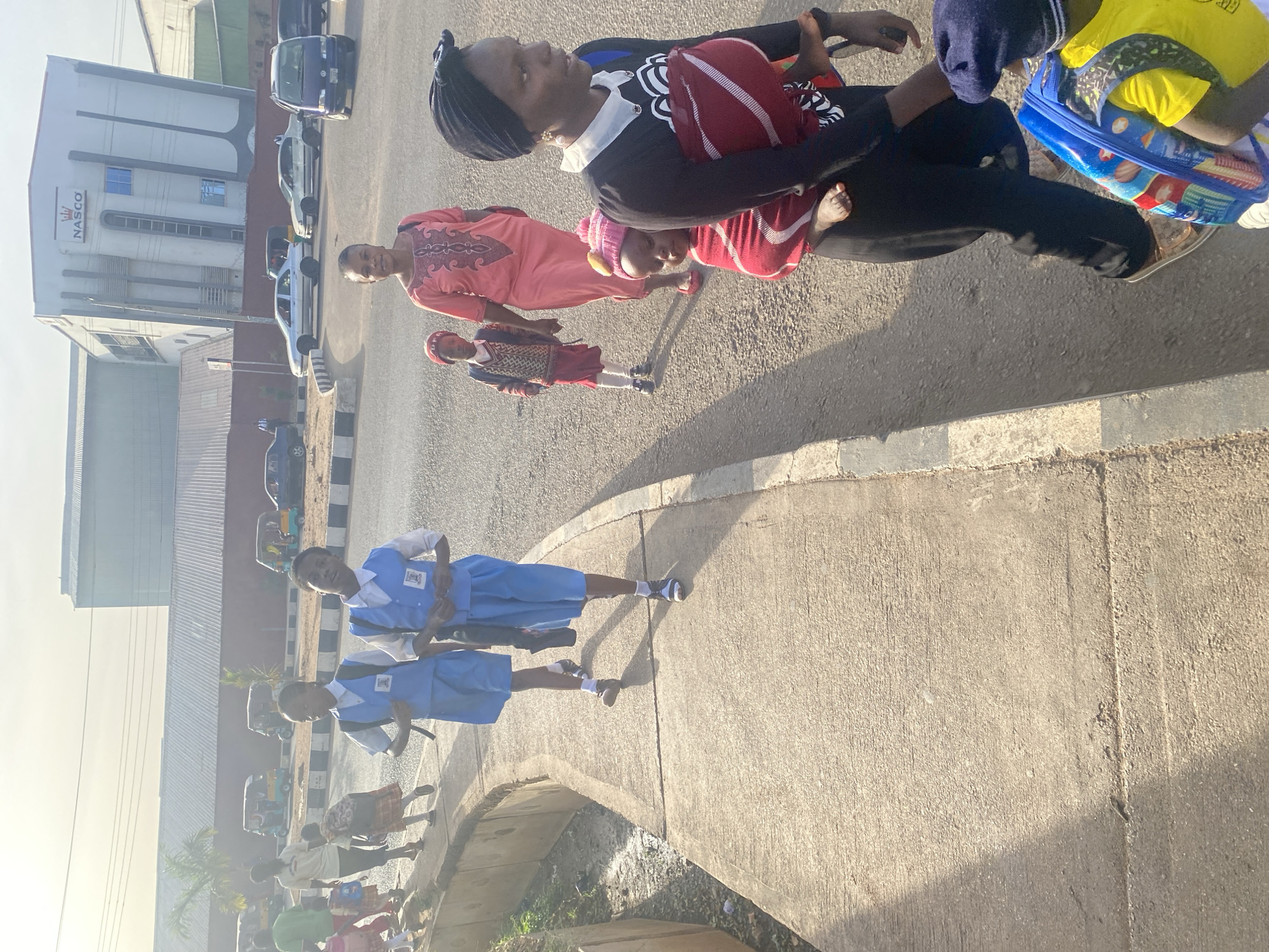 The image captures a vibrant, sunny street scene in an urban area, likely in Jos, Nigeria. The photograph is taken from a slightly elevated perspective, looking down at a concrete sidewalk and an asphalt road.
In the foreground, a group of pedestrians is visible. Prominently, two young girls wearing matching light blue and white school uniforms, white socks, and dark shoes walk towards the viewer on the sidewalk. Their uniforms feature a small, dark emblem on the chest.
Further into the mid-ground, another cluster of individuals is seen. A woman in a dark dress with a white collar and a patterned design, along with a dark headscarf, holds a baby or very young child wrapped in a red and white striped blanket. Beside her, a woman in a bright pink dress with dark embroidery stands, offering a slight smile. A young boy in a patterned sweater and red shorts is also part of this group, along with another small child in a red and pink striped outfit and a pink hat. To the right, another child is visible, carrying a blue and yellow backpack.
The background is dominated by a large, multi-story white building with "NASCO" clearly written in dark, capital letters on its facade, accompanied by a crown logo. Several cars of various models and colors are parked or moving on the road in front of the building. In the distance, more buildings and other vehicles, including what appear to be auto-rickshaws, are visible.
The strong, distinct shadows cast by the people and structures indicate clear skies and intense sunlight, suggesting either a morning or late afternoon setting. The scene depicts everyday life with people walking along the street, possibly commuting to or from school and work.