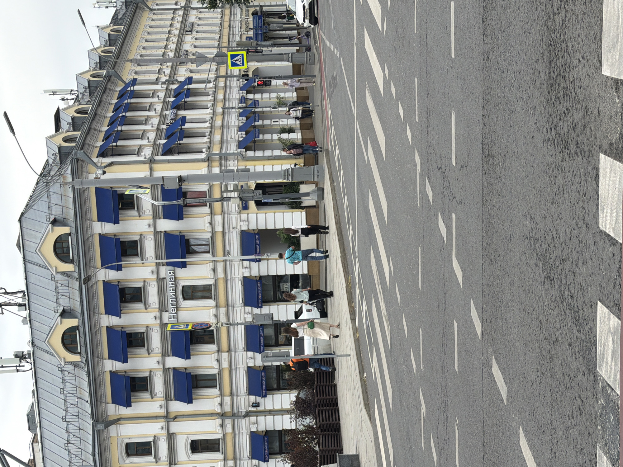 The image depicts a street scene in Moscow, Russia, likely near Neglinnaya Street, as indicated by the Cyrillic signage on the building. A multi-story building with a yellow facade and blue awnings over the windows dominates the background. The building appears to be commercial, with storefronts on the ground floor. Several individuals are visible on the sidewalk, including people walking and standing. A white car is parked on the street. The foreground features a pedestrian crossing marked with white stripes on the asphalt. Traffic lights and street lamps are also present, suggesting an urban environment. The sky is overcast, indicating a cloudy day.