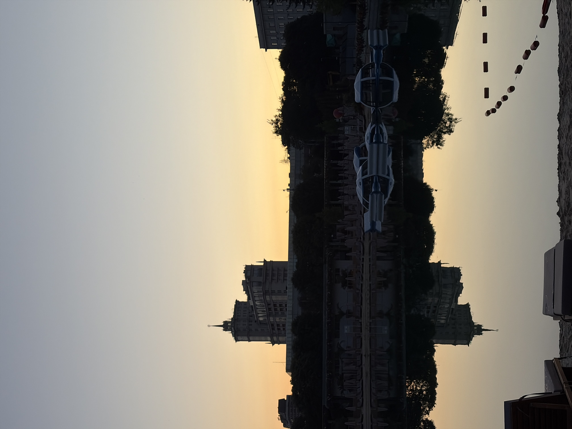 This is an image of a lake in Gorky Park, Moscow, Russia, taken at sunset. The sky is a gradient of pale blue to orange, with the sun low on the horizon. In the background, silhouetted against the sky, are several buildings, including a tall, ornate structure that appears to be a hotel or government building. There is also a line of trees and some smaller buildings. In the foreground, the lake is calm and reflects the sky and buildings. Several white, pod-like floating structures are on the water, and a rope with buoys marks off a section of the lake. The beach area in front of the lake has loungers and umbrellas, some of which are folded. The overall impression is one of tranquility and natural beauty within an urban setting.