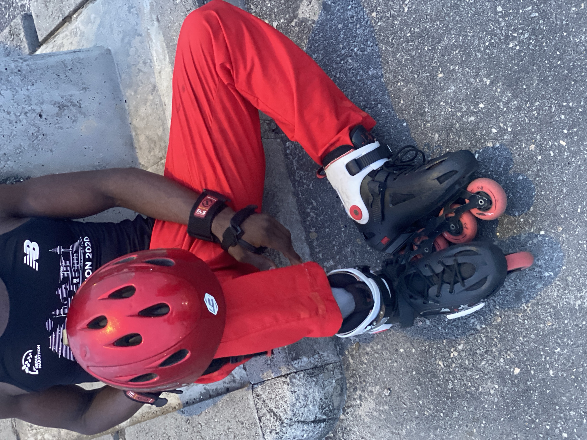 The image shows a person sitting outdoors on a concrete surface, possibly a step or low wall. The person is wearing red pants, a black tank top with a New Balance logo and a "SEOUL MARATHON 2020" graphic, black inline skates with white and red details, a red helmet, and wrist guards. The location is not explicitly identified as Sangotedo, Nigeria, but given the clothing and skin tone of the subject, it may be accurate.