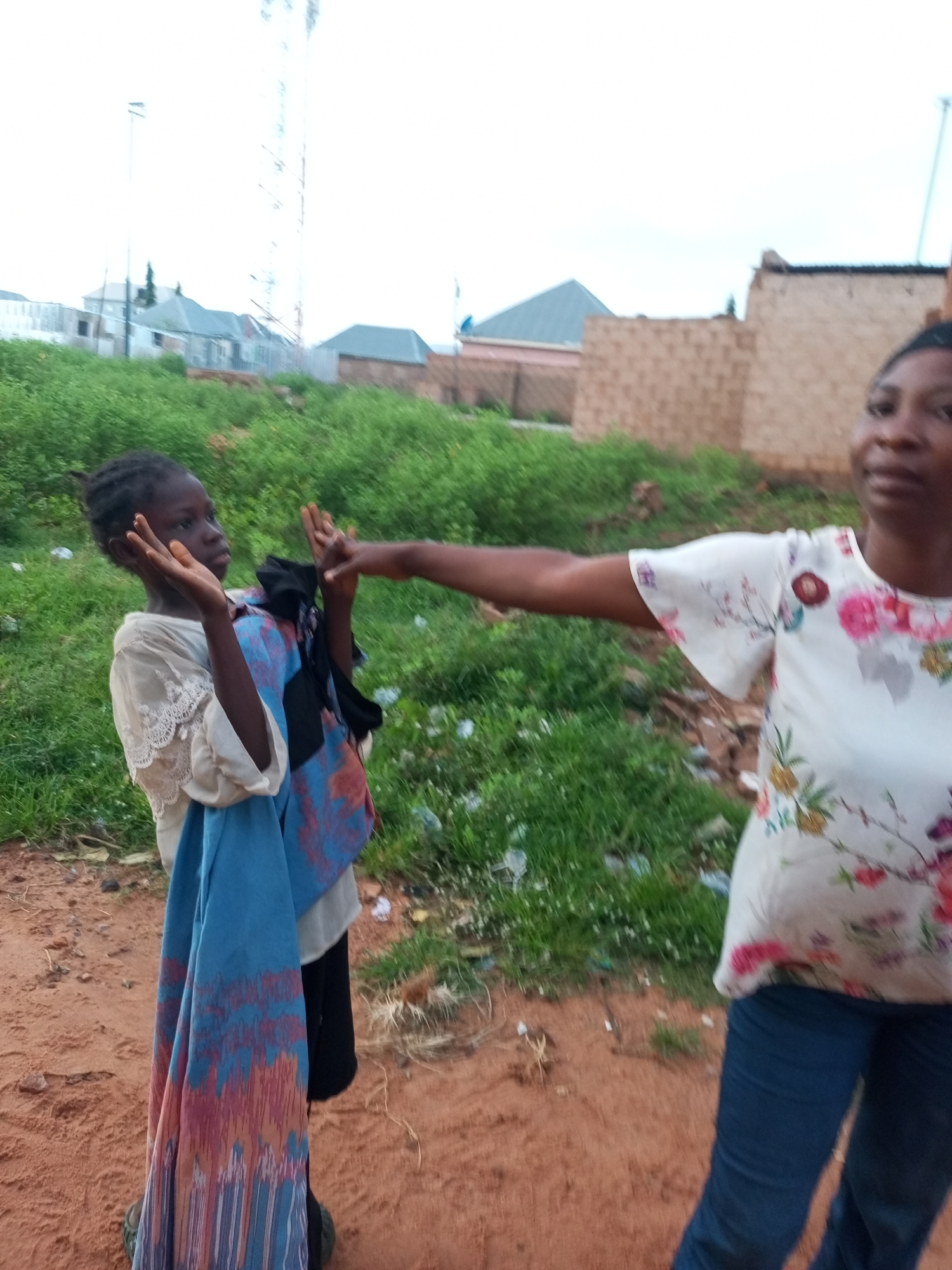 In the city of Tempure, Nigeria, a young girl stands on a dirt path, her hands raised as if in greeting or surprise. She is wearing a light-colored shirt with intricate lace detailing around the collar and sleeves, and a blue and red patterned fabric is draped over her left arm. To her right, a woman in a white floral-print shirt and blue jeans extends her right arm towards the girl, her hand also raised. The background features a grassy slope with scattered debris, and a few buildings and a tall communication tower are visible in the distance under a cloudy sky.