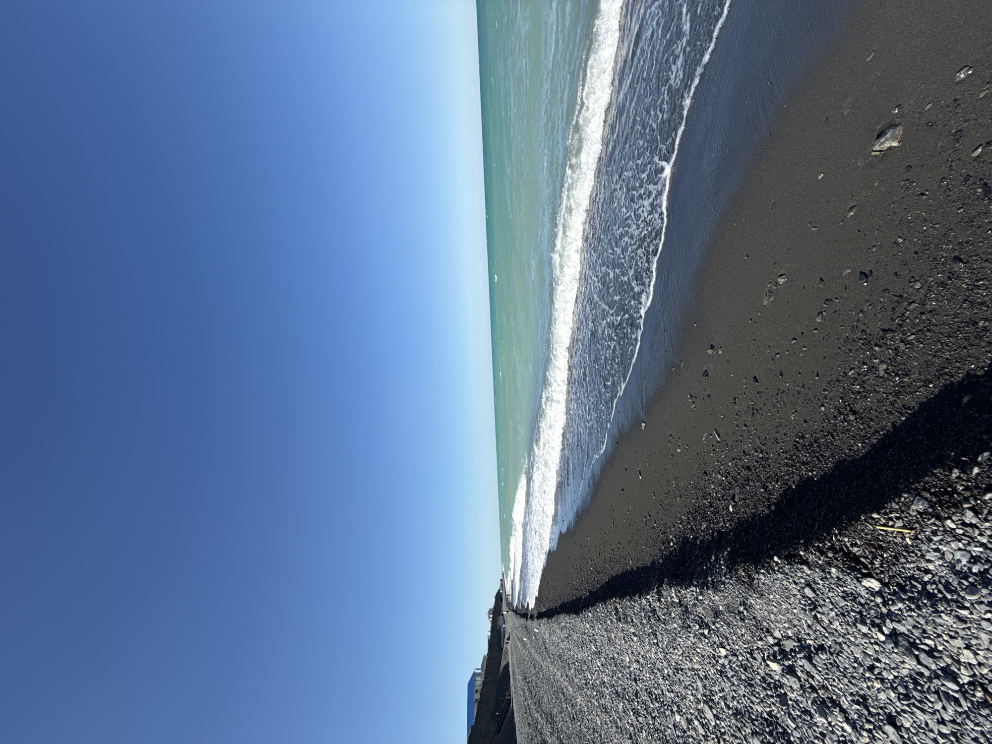A wide-angle, eye-level shot captures a coastal scene on a clear, sunny day, with a brilliant blue sky dominating the upper portion of the frame. The foreground features a stark contrast between two types of beach: a dense, grey pebble beach on the left, and a darker, black sand beach on the right. A distinct shadow line separates the two textures. The black sand beach meets a turquoise-colored sea with gentle, white-capped waves. In the distance, along the edge of the pebble beach, a line of buildings with blue and white roofs is visible, nestled against a dark, sloped embankment. A few scattered figures are present on the pebble beach pathway in the mid-ground, appearing small and distant. The overall impression is one of a serene, unpopulated coastline. The specific location is the beach in Ақши, Kazakhstan.