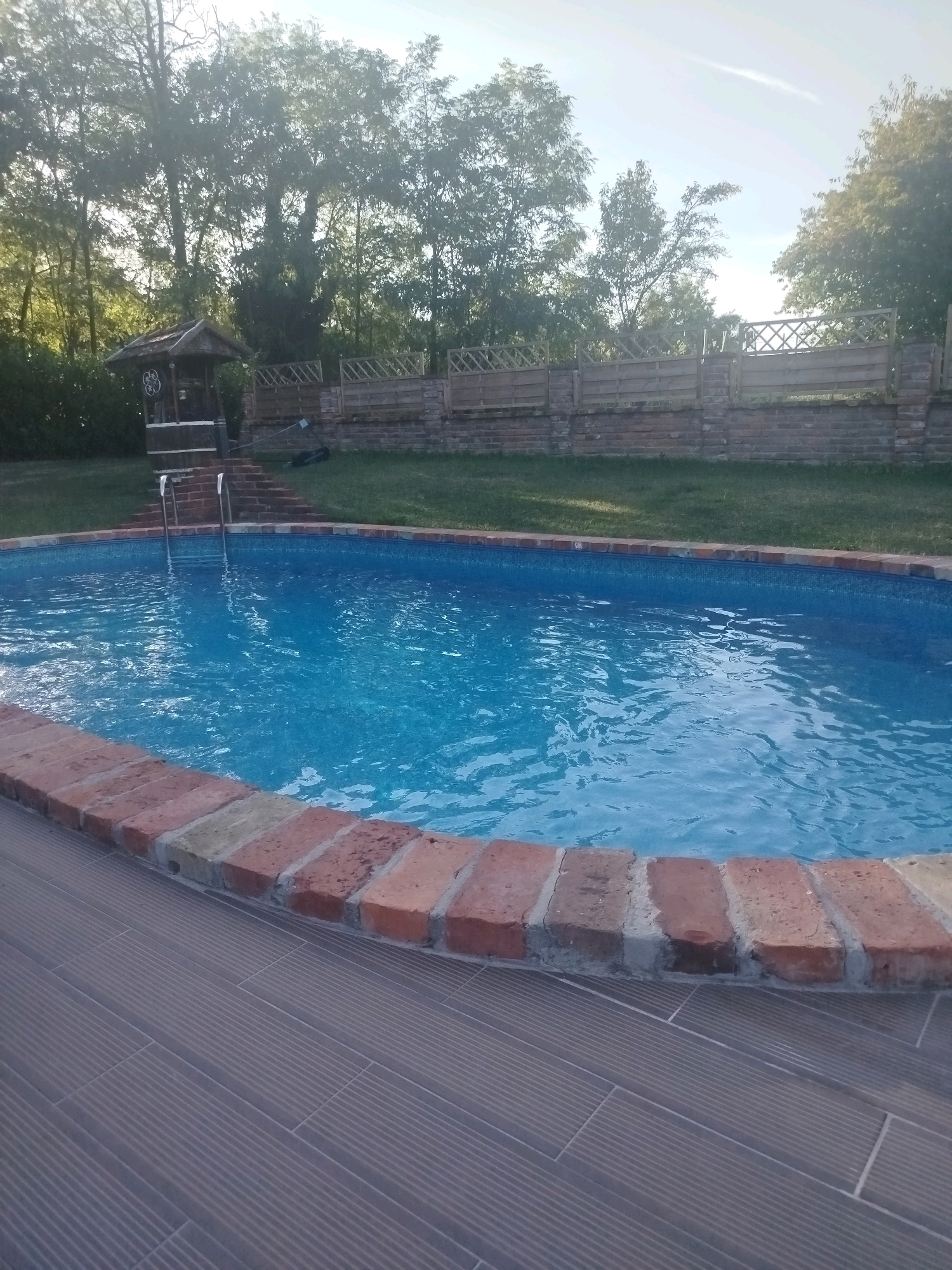 An oval-shaped swimming pool filled with clear blue water is centrally located in the foreground of the image. The pool is bordered by a low wall constructed of reddish-brown bricks. Beyond the pool, a paved area with dark grey tiles extends to the left. To the right of the pool, a lush green lawn is visible. In the background, several mature trees with dense green foliage create a natural backdrop. To the left of the pool, a rustic wooden structure resembling a well or a pump house stands, with stairs leading up to it. A wooden fence with diamond-shaped patterns separates the lawn area from a brick wall further in the background. The sky above is a pale blue with a hint of haze, suggesting it is daytime. The image appears to be taken in a backyard or garden setting, possibly in Brckovljani, Croatia, given the context provided. There are no people or animals visible in the scene.