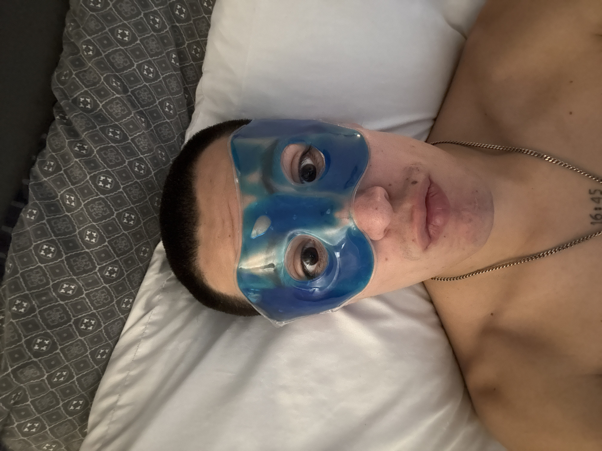 A person is lying on a bed with a blue gel eye mask. The person has short, dark hair and a silver chain necklace. A tattoo with the numbers "16:45" is visible on the chest. The person is resting on a white pillow with a grey patterned blanket visible on the left. It is not possible to determine the specific location in San Bernardino Tlaxcalancingo, Mexico, from this image.