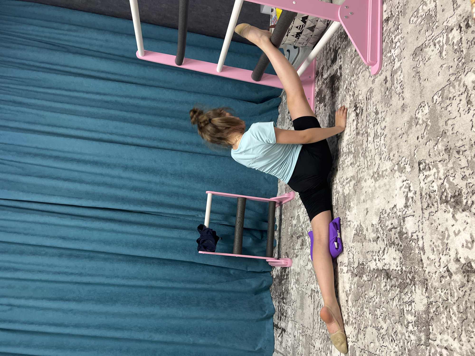 A young girl, with light brown hair styled in a bun, is performing a split in an indoor setting. Her left foot is resting on a horizontal rung of a pink and white children's wall gym. Her right leg extends downwards, supported by a purple cushion placed between her leg and the patterned gray and white floor. She's wearing a light blue t-shirt and black shorts. Her right hand is on the floor for balance.
The wall gym, which is primarily pink and white, is affixed to a wall with a patterned surface. To the left, a turquoise curtain hangs vertically. A smaller, identical pink and white gym apparatus is partially visible to the left, with a dark-colored item on one of its rungs.
The location is identified as Chaikovsky, Russia, based on image metadata (not visible).