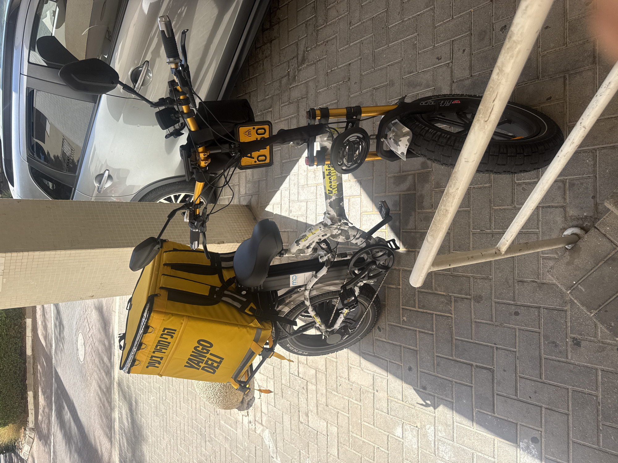 A yellow delivery bag branded "YANGO DELI" with Hebrew text is mounted on the rear rack of a black and white camouflage-patterned electric bicycle. The bicycle has yellow handlebars and front suspension. A yellow license plate with black characters "A 0150" is attached to the handlebars. The bicycle is parked on a herringbone-patterned brick sidewalk next to a building with a tiled facade and a silver car. The scene is outdoors, with sunlight casting shadows. The image was taken in Herzliya, Israel.
