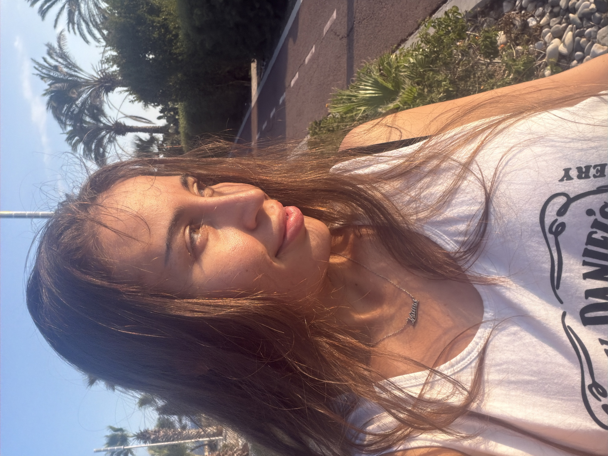 In the city of Meneou, Cyprus, a young woman with long, brown hair is captured in a close-up, head-and-shoulders shot. She is wearing a white tank top with a black strap visible over her shoulder. A silver necklace with a nameplate spelling "Meme" rests on her collarbone. The woman is looking to her right, with the sun casting a warm glow on her face, highlighting her cheekbones and lips. Her hair is gently blowing in the breeze. Behind her, a paved pathway with white markings is visible, bordered by lush green bushes and palm trees under a clear blue sky. The ground to the right of the pathway is covered in small, light-colored stones. The overall scene suggests an outdoor setting in a sunny, possibly coastal, environment.