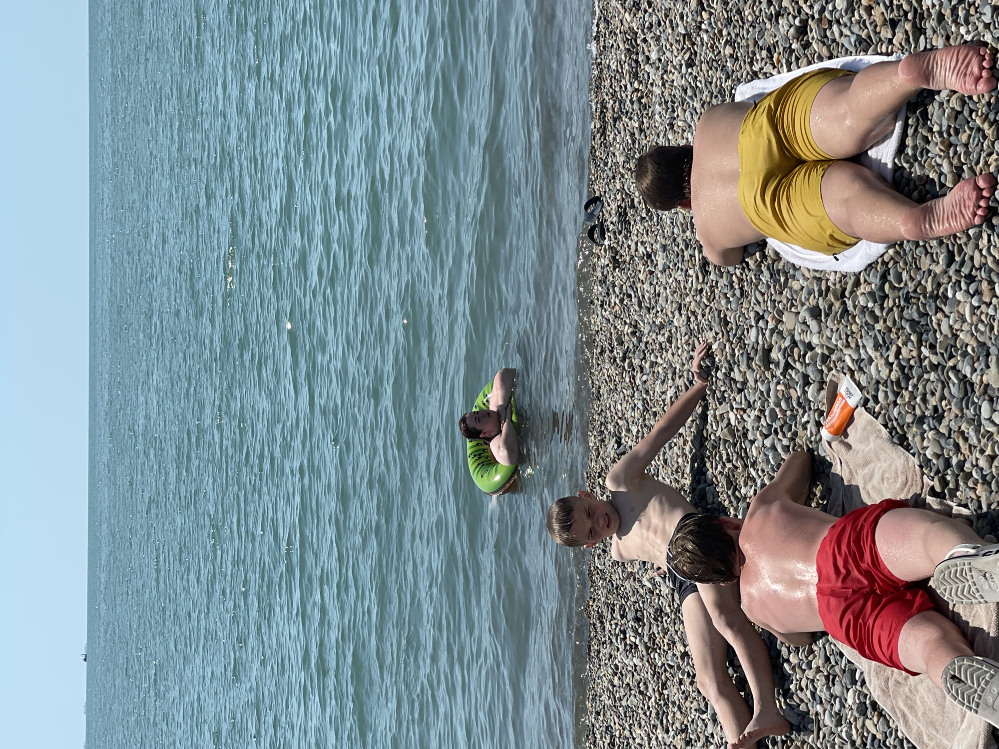 In Sochi, Russia, on a sun-drenched day, three young males are present at a pebble beach adjacent to the Black Sea. The water is a deep blue, with gentle ripples and a clear horizon.
The first male, lying on his back, is positioned on the right side of the frame. He is wearing yellow swimming shorts and resting on a white towel. His back and legs are glistening with what appears to be sunscreen or water, indicating recent exposure to either.
In the middle of the frame, closer to the water's edge, another male is lying on his stomach. He is wearing red swimming shorts and is also on a towel, which is light beige. His back is also wet and shiny, reflecting the sunlight. An orange bottle, likely sunscreen, is placed on the towel beside him.
To the left of the frame and closer to the water, a third male is in a reclined position on the pebbles, facing towards the viewer with a slight grimace. He is wearing dark swimming shorts and his torso is bare.
Further into the water, a fourth individual is seen relaxing in a bright green inflatable ring. They are in a seated position, leaning back against the ring with their arms resting on it. The water is shallow around them. In the distance, several ships are visible on the horizon line, against a pale blue sky. The entire scene is illuminated by bright sunlight.