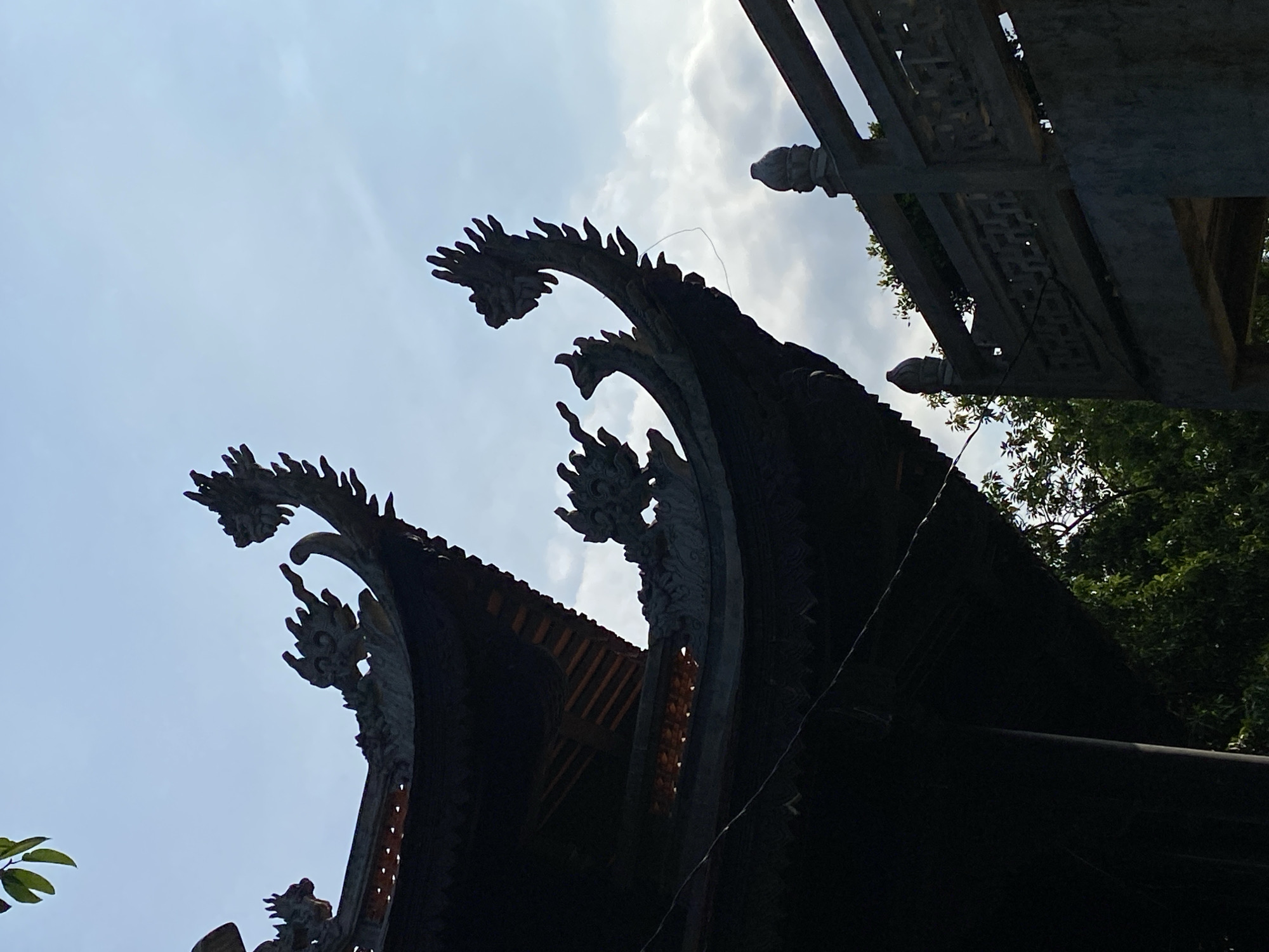 The image shows the ornate, dark-tiled roof of a traditional Vietnamese building, possibly a temple or pagoda, with stylized dragon motifs adorning its eaves. The architecture is silhouetted against a bright, partly cloudy sky. In the foreground to the right, a section of a stone balustrade with decorative carvings is visible, along with the top of a stone pillar. Lush green foliage from surrounding trees peeks out from behind the structures. The overall impression is one of traditional East Asian architectural beauty under a bright sky.