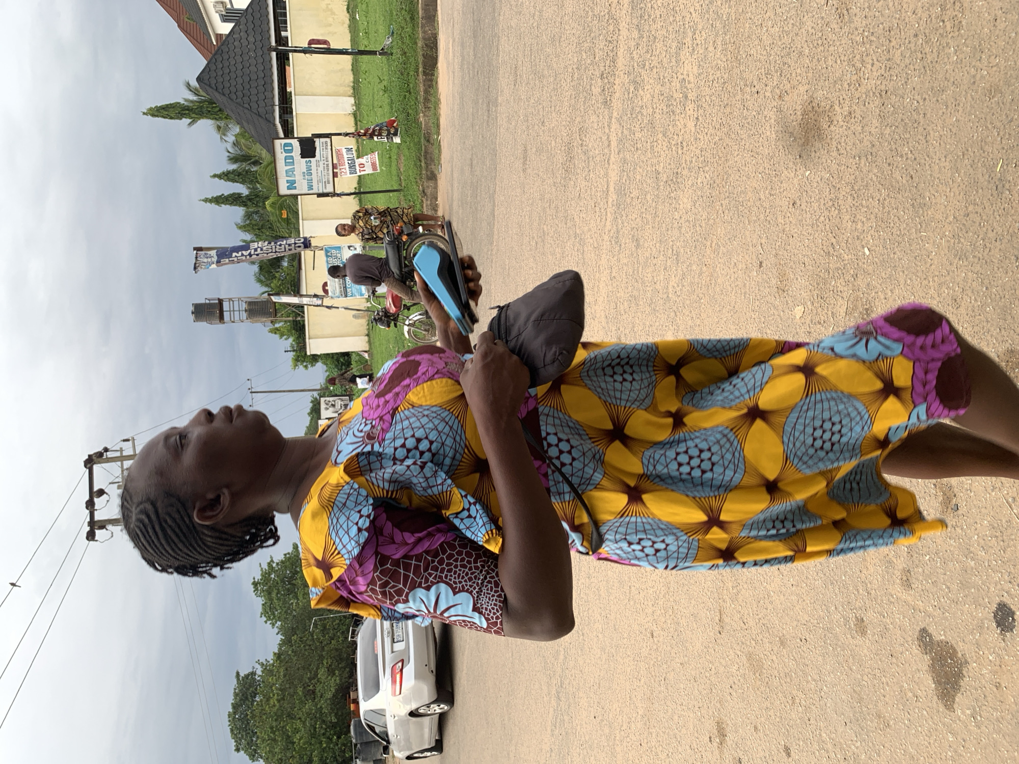 A medium shot captures a Nigerian woman from the waist up, walking on a sandy road in Abuja, Nigeria. She wears a vibrant, patterned yellow, blue, and purple dress with puffy sleeves. Her dark braided hair is pulled back. In her right hand, she holds a dark gray cloth pouch, and in her left hand, she grips a blue card reader and a smartphone.
In the background, a silver sedan drives away on the road. Further back, a red motorcycle is parked, with a man in a brown shirt and a woman in a patterned dress standing beside it. A billboard with the text "NADO" and "AND WILLOWS" is visible, along with other signs and a residential building with a dark, conical roof. A water tower stands tall behind the yellow wall. The sky is overcast.