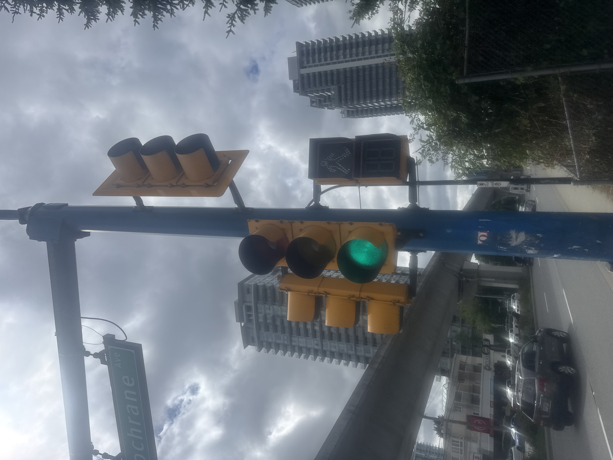 The image depicts a street scene in Burnaby, Canada, under a cloudy sky. A traffic light with a green light illuminated is prominently featured, along with a pedestrian signal displaying a walking figure and the number "20". Above, a street sign reads "Cochrane Ave". In the background, modern high-rise buildings and an elevated transit line are visible. Several vehicles are present on the road, including a dark SUV in the foreground. Vegetation, including trees and bushes, lines the side of the road, and a chain-link fence can be seen on the right. The overall atmosphere is urban, with evidence of transportation infrastructure and residential buildings.