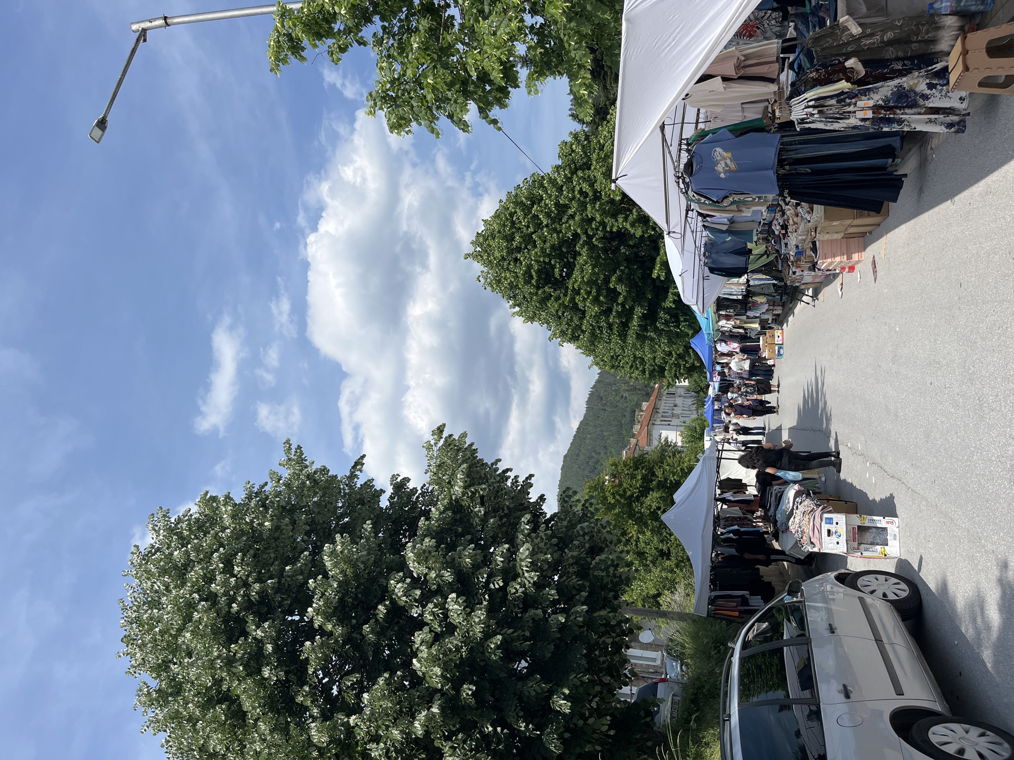 This detailed description portrays an outdoor street market in Madan, Bulgaria, observed on a bright, partly cloudy day.
The scene is dominated by a paved road that serves as the market aisle, lined on both sides with numerous vendor stalls operating under white and blue canopy tents. The stalls predominantly display various articles of clothing, including t-shirts, dresses, skirts, jeans, and other garments, hung on racks or neatly folded on tables and in cardboard boxes. A blue t-shirt with a graphic print is prominently displayed on a hanger on the right side, along with several floral-patterned dresses and denim items. A small brown plastic stool and a blue plastic water bottle are visible on the ground near a stall on the right.
Approximately 15 to 20 individuals are present throughout the market. Most are engaged in browsing the merchandise at the stalls or walking along the thoroughfare. On the left side, a person in dark clothing stands near a display of folded clothes. Further down the street, groups of people can be seen meandering between the stalls.
On the far left, a silver minivan or large station wagon is parked, facing away from the camera. Its license plate is partially visible, starting with "CB 777 EK". Large, mature trees with dense green foliage, casting shadows on the road, frame the left side of the image and extend into the background.
The background features additional lush green trees, older multi-story buildings with red-tiled roofs, and distant, forested hills that rise against the horizon. A modern grey street lamp with an LED fixture extends over the road from the upper right. The sky is a clear blue with scattered white cumulus clouds, indicating fair weather conditions and bright daylight. Shadows cast by the trees and stalls suggest the sun is high, likely indicating late morning or early afternoon.