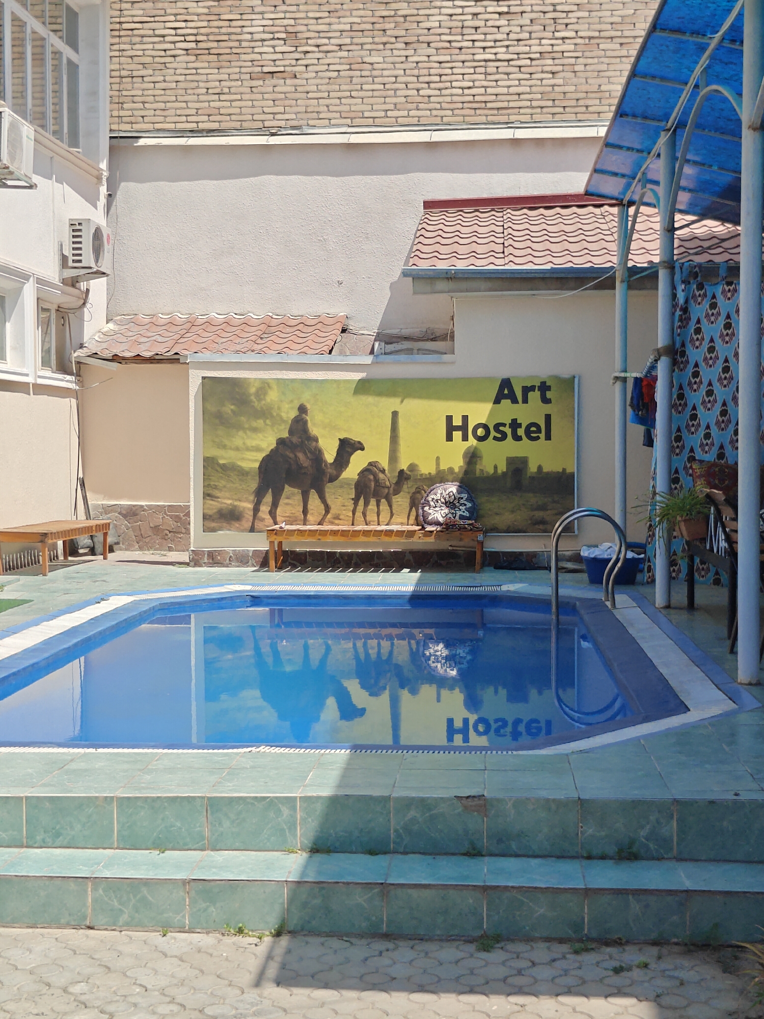 The image shows an outdoor swimming pool area in front of a building. The pool is rectangular and filled with blue water. The surrounding area is paved with light-colored tiles. To the left of the pool, there is a wooden bench. Behind the pool, there is a large mural depicting camels and a desert landscape with buildings. The mural also has the text "Art Hostel" on it. To the right of the pool, there is a blue canopy supported by poles, and patterned curtains. The building in the background has a brick facade. The scene appears to be in a sunny, outdoor location. This is the Art Hostel in Tashkent, Uzbekistan.