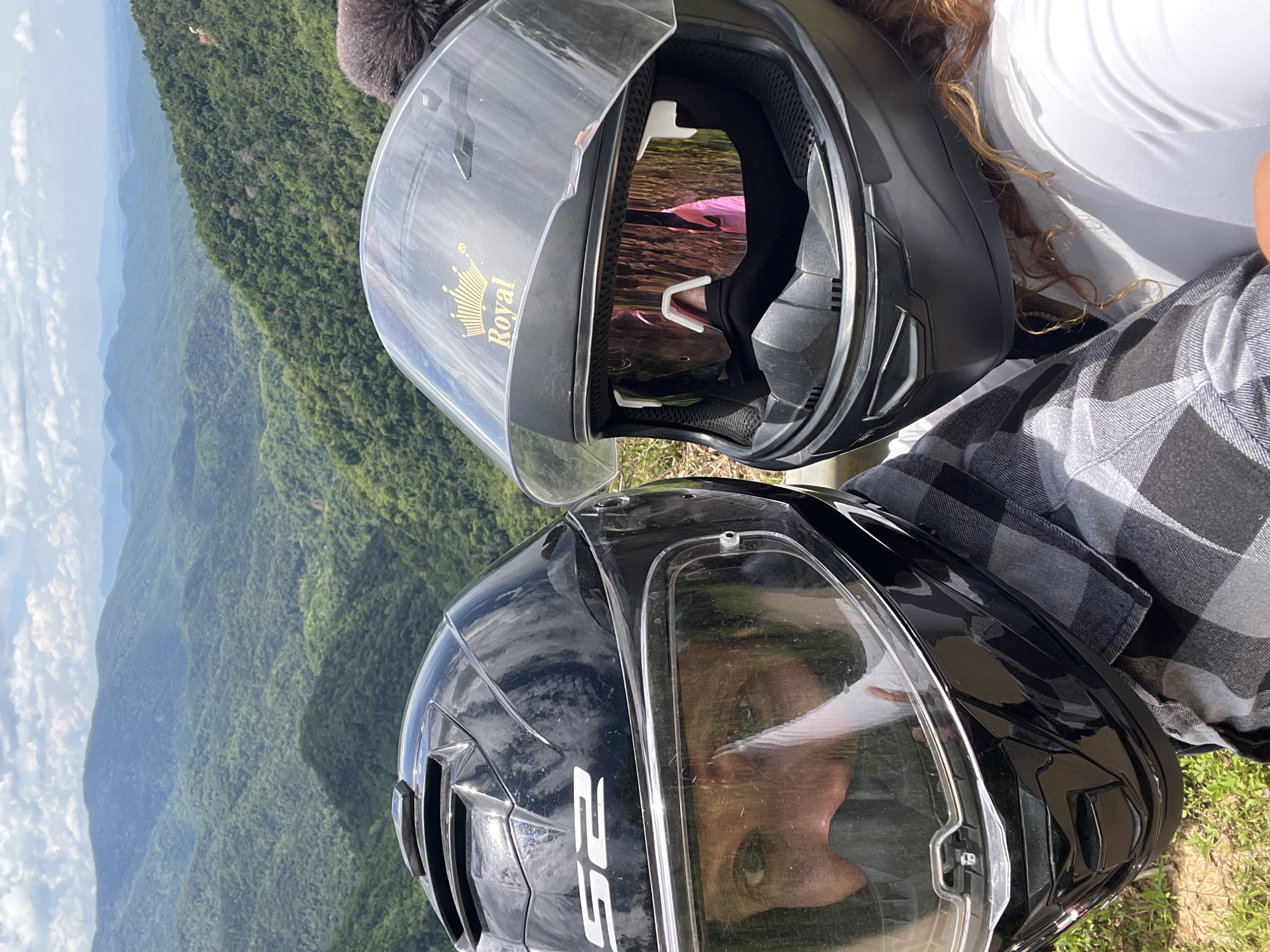A selfie taken by two individuals wearing motorcycle helmets is captured against a backdrop of lush, verdant mountains under a cloudy sky. The person on the left, wearing a black helmet with "LS2" branding, has their eyes visible through the visor. The person on the right wears a black helmet with a gold "Royal" crown decal on the top and mirrored goggles. The helmets obscure the faces of the individuals, with only the eyes and parts of their faces visible. The background shows a panoramic view of rolling green hills and mountains, characteristic of a mountainous region like Son Dien, Vietnam. The lighting suggests it is daytime.