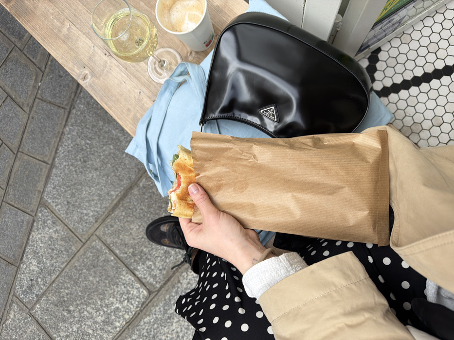 The image provides a first-person, overhead perspective of a person seated at a wooden table in an environment consistent with an outdoor or semi-outdoor cafe in Paris, France. No specific landmarks are visible to pinpoint an exact location within the city.
The person's left hand, adorned with a subtle wrist tattoo, holds a partially eaten baguette sandwich, displaying red (tomato) and green (greens) fillings, inside a brown paper bag. Visible attire includes the sleeve of a tan trench coat, a white ribbed cuff, and a black garment with white polka dots on the lap. A black shoe is visible on the grey cobblestone pavement to the left.
On the wooden table surface, a clear stemmed wine glass containing a pale yellow liquid (suggesting white wine) and a white paper cup with a foamy light brown beverage (likely coffee) are present. A light blue fabric item, possibly a tote bag, is draped on the table. Adjacent to these items is a black, shiny Prada handbag, featuring a distinct silver triangular logo. The floor visible below the table is covered with a white and black hexagonal tile pattern.