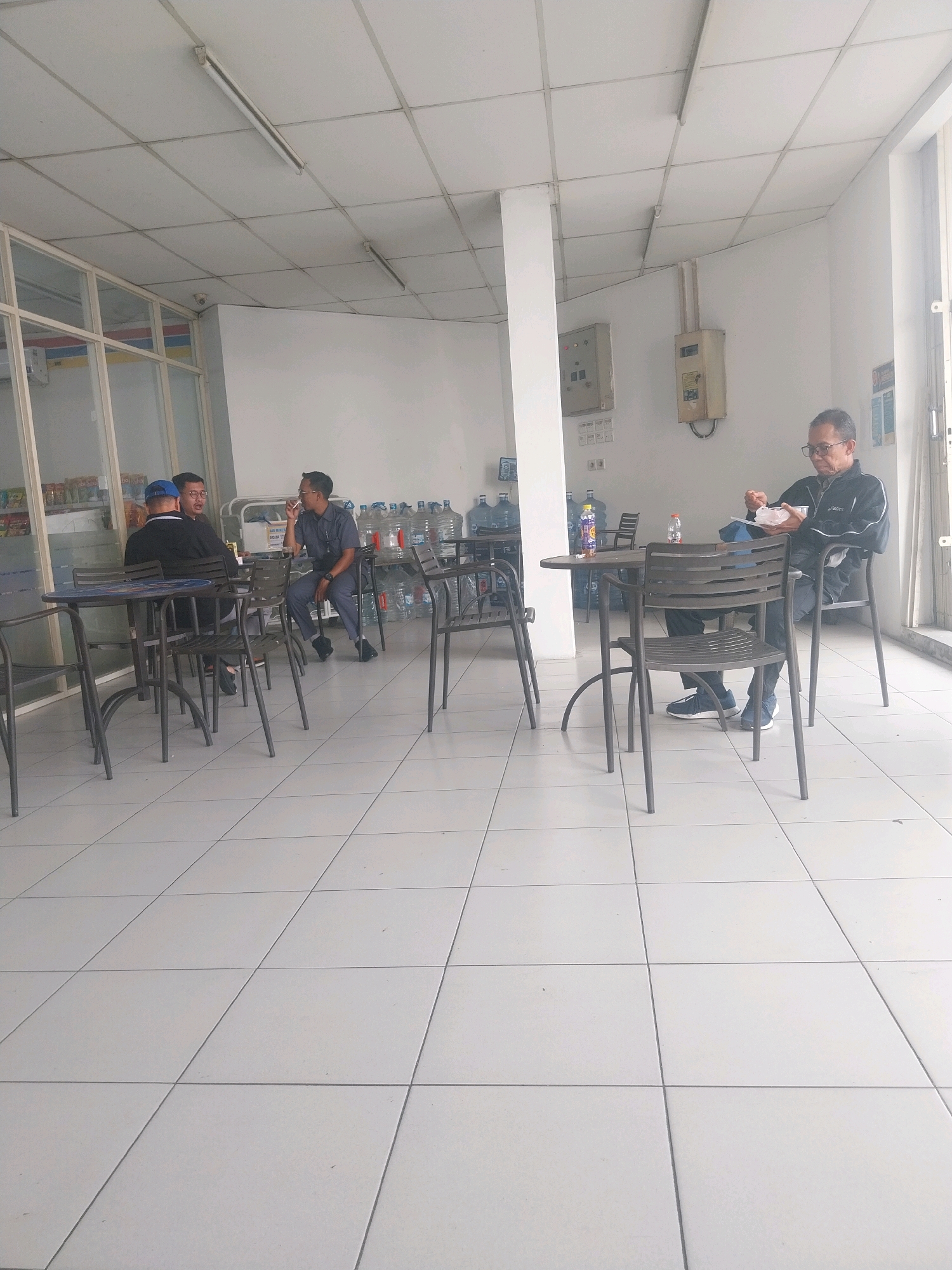 The image depicts an indoor cafe or rest area, likely a stopover point along a highway or a rest area within a facility in Karawang, Indonesia. The environment is characterized by a tiled floor and a suspended ceiling with fluorescent lighting.
There are four men present. Two men are seated at a table on the left, engaged in conversation. One of them wears a blue baseball cap and a dark jacket, while the other is in a dark shirt and glasses. In the background, a third man in a dark uniform and glasses is seated at another table, seemingly on his phone or engaged in thought. On the right, a fourth man, also in dark attire and glasses, is seated at a table, eating from a bowl with a spoon.
The cafe area is furnished with simple metal tables and chairs. In the background, several large bottles of drinking water are visible, along with shelves that appear to stock snacks or other convenience items. A window or glass partition is present on the left side of the image, offering a glimpse of what might be a store or more public area. Electrical conduits and utility boxes are mounted on the wall in the upper right portion of the frame, suggesting a functional, rather than purely aesthetic, space. The overall impression is that of a utilitarian rest stop designed for convenience.