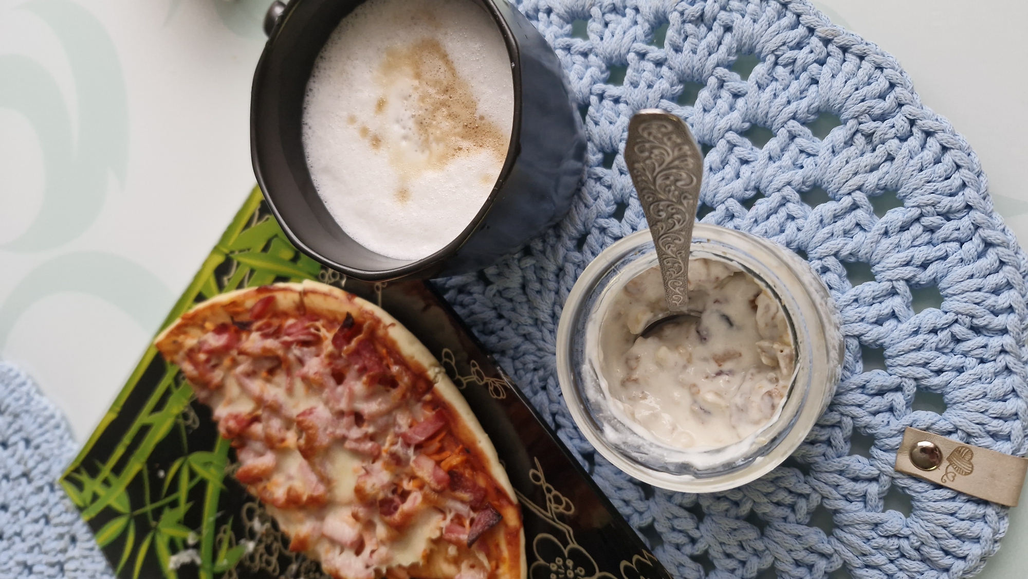 This overhead shot showcases a casual meal laid out on a light surface, likely a table, against a textured, light blue crocheted placemat. A dark gray or black mug, positioned centrally, holds a beverage with a thick layer of white foam, suggesting coffee like a latte or cappuccino, with some light brown specks on top.
To the left, a slice of pizza rests on a rectangular serving plate or tray. The plate is black with a vibrant green bamboo leaf pattern. The pizza slice is topped generously with melted cheese and diced ham or bacon, exhibiting a golden-brown crust.
On the right side, a clear glass jar contains a creamy mixture, possibly overnight oats, yogurt with granola, or a similar breakfast item. A silver spoon with an ornate, antique-style handle is partially submerged in the contents of the jar.
The light blue crocheted item, which functions as a placemat or large coaster, features an intricate, openwork pattern. In the lower right corner of the placemat, a small, light brown leather-like tag is attached with a silver rivet. This tag bears an embossed design that appears to be a stylized yarn ball or a knitting motif, suggesting a handmade touch.
The arrangement implies a relaxed breakfast or brunch setting, combining savory and possibly sweet elements with a warm drink. There are no people visible, and no specific time of day or weather conditions can be discerned from the image itself. The location context provided for this media file is Otchor, Russia. No discernible text is present within the image apart from the embossed design on the tag.