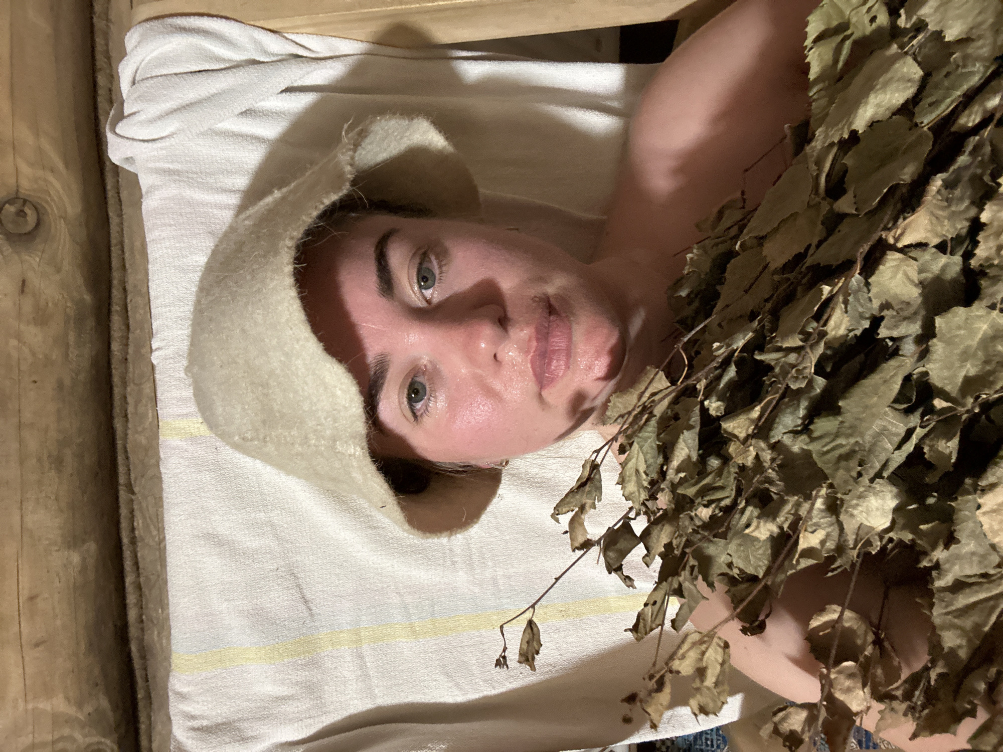 A woman with fair skin, wearing a felt sauna hat, lies on a striped towel on what appears to be a wooden sauna bench. She holds a bundle of dried birch branches (a "venik") close to her body. Her skin is moist, suggesting she is in a sauna. The location may be Уральское, Russia, based on image origin.
