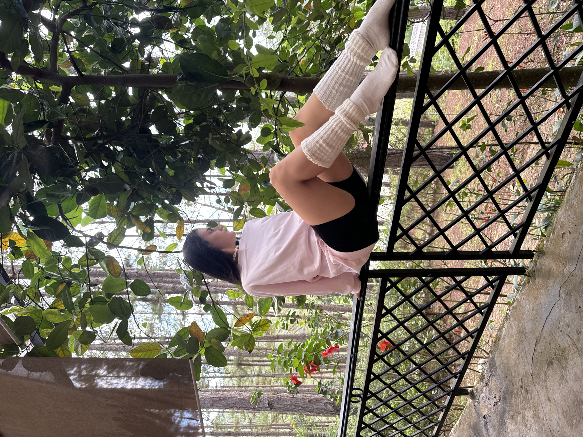 A woman is reclining on a dark metal railing, positioned between the viewer and a background of dense foliage and a wooded area. She is wearing black shorts, a light pink long-sleeved shirt, and white leg warmers. The setting is verdant, with trees visible both above and in the distance behind the railing. Based on the architectural style and landscape, the location is very likely Da Lat, Vietnam.