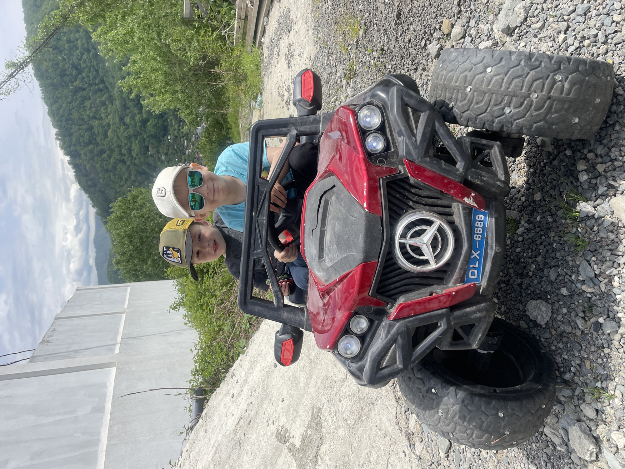 Two young boys are sitting in a red and black toy Mercedes-Benz-style off-road vehicle. The vehicle has a license plate that reads "DLX-6688." The tire closest to the viewer has metal studs in the treads. The background features a landscape with a concrete wall, green trees, and a distant mountain range, consistent with the Baranovka, Russia, area. The boys appear to be posing for a photograph. One boy is wearing a blue shirt and sunglasses, the other a hat with a cartoon image. The vehicle is situated on a rough, gravel-covered surface.