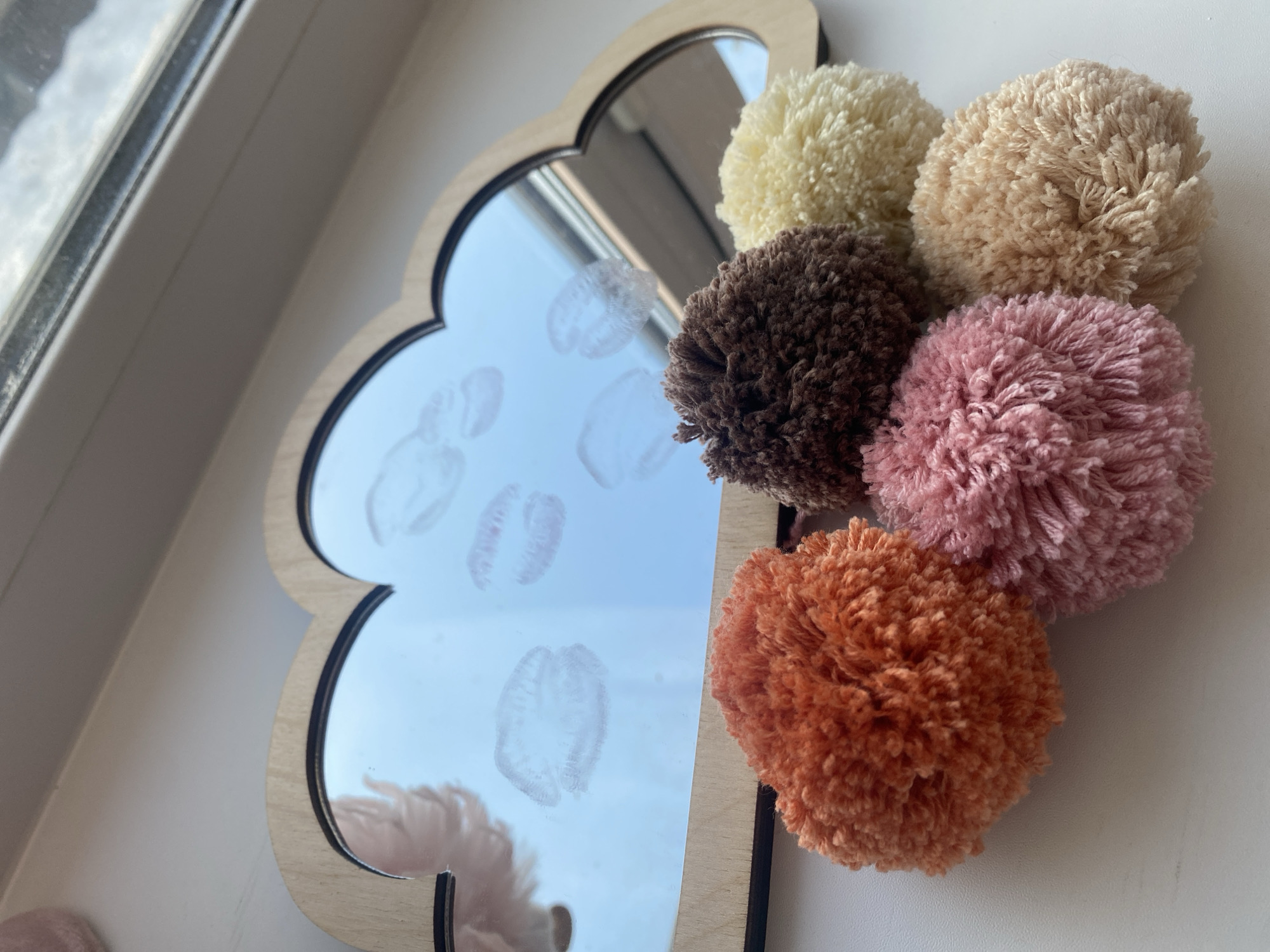 A close-up shot shows a cloud-shaped mirror with a light wood frame, decorated with six colorful pom-poms. The mirror reflects a window and the pale sky outside. Several faded lip prints are visible on the mirror's surface. The pom-poms are arranged in a cluster, with colors including yellow, beige, brown, rose, and orange. The background is a white wall. It's not possible to know where this photo was taken, and there is no indication that it was taken in Izhevsk, Russia.