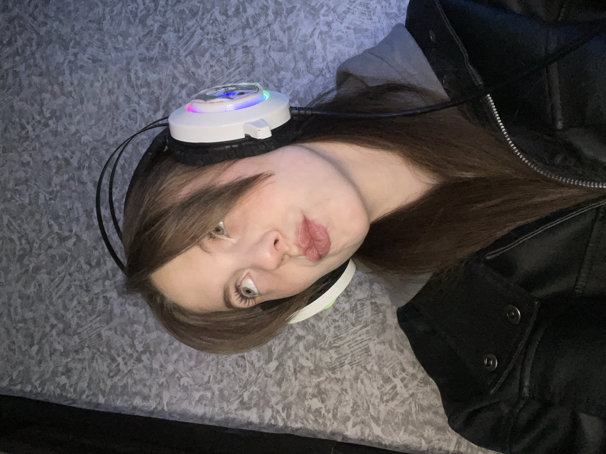 A woman with brown hair and fair skin is lying down wearing headphones with a white, lighted top and black ear covers. She is wearing a dark jacket. The background appears to be a gray patterned surface. The location is unknown and is not discernible from the image. The specified location cannot be confirmed.