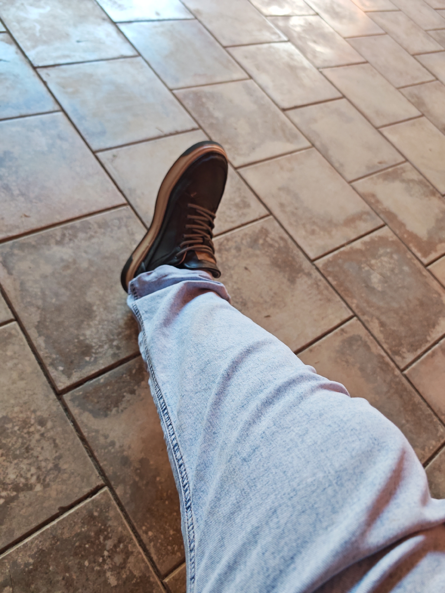 The image shows a person's leg and foot, clad in light blue jeans and a dark brown boot, resting on a tiled floor. The tiles are rectangular and arranged in a staggered pattern. The lighting suggests an indoor or shaded outdoor environment. The image does not provide any specific visual cues to identify the location as Mayrtup, Russia.