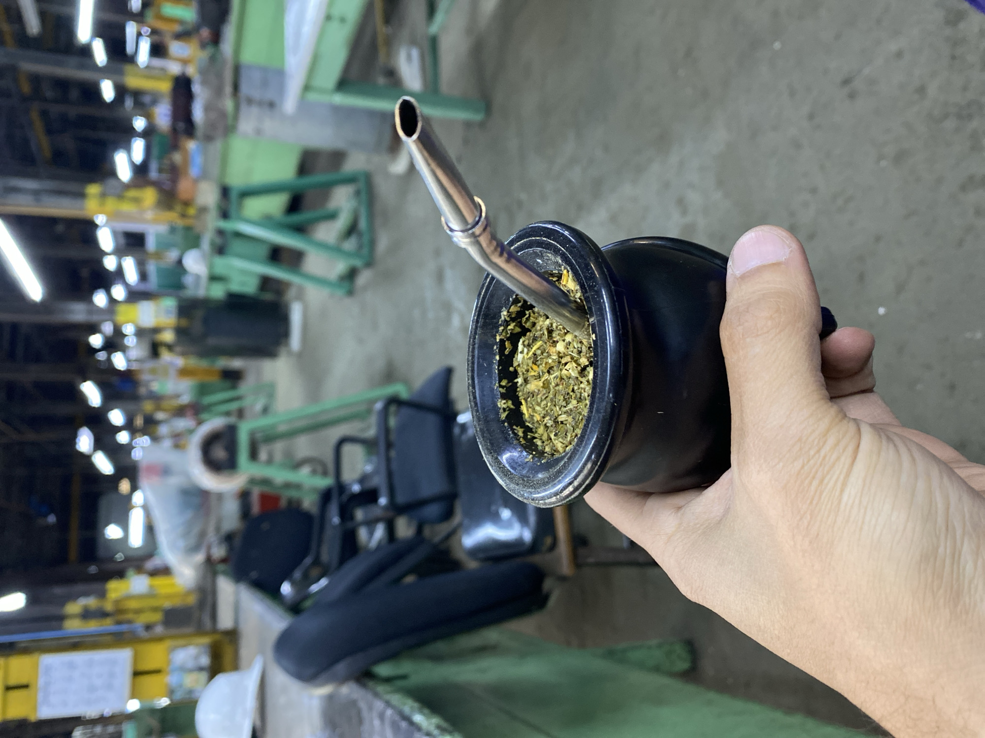 A person holds a black mate gourd with a metal bombilla (straw) inserted. The gourd is filled with yerba mate. The person's hand is light-skinned. The background shows an industrial setting, possibly a factory or workshop. There are metal tables and chairs, along with overhead lighting. The scene does not explicitly confirm that this is in Buenos Aires, Argentina.