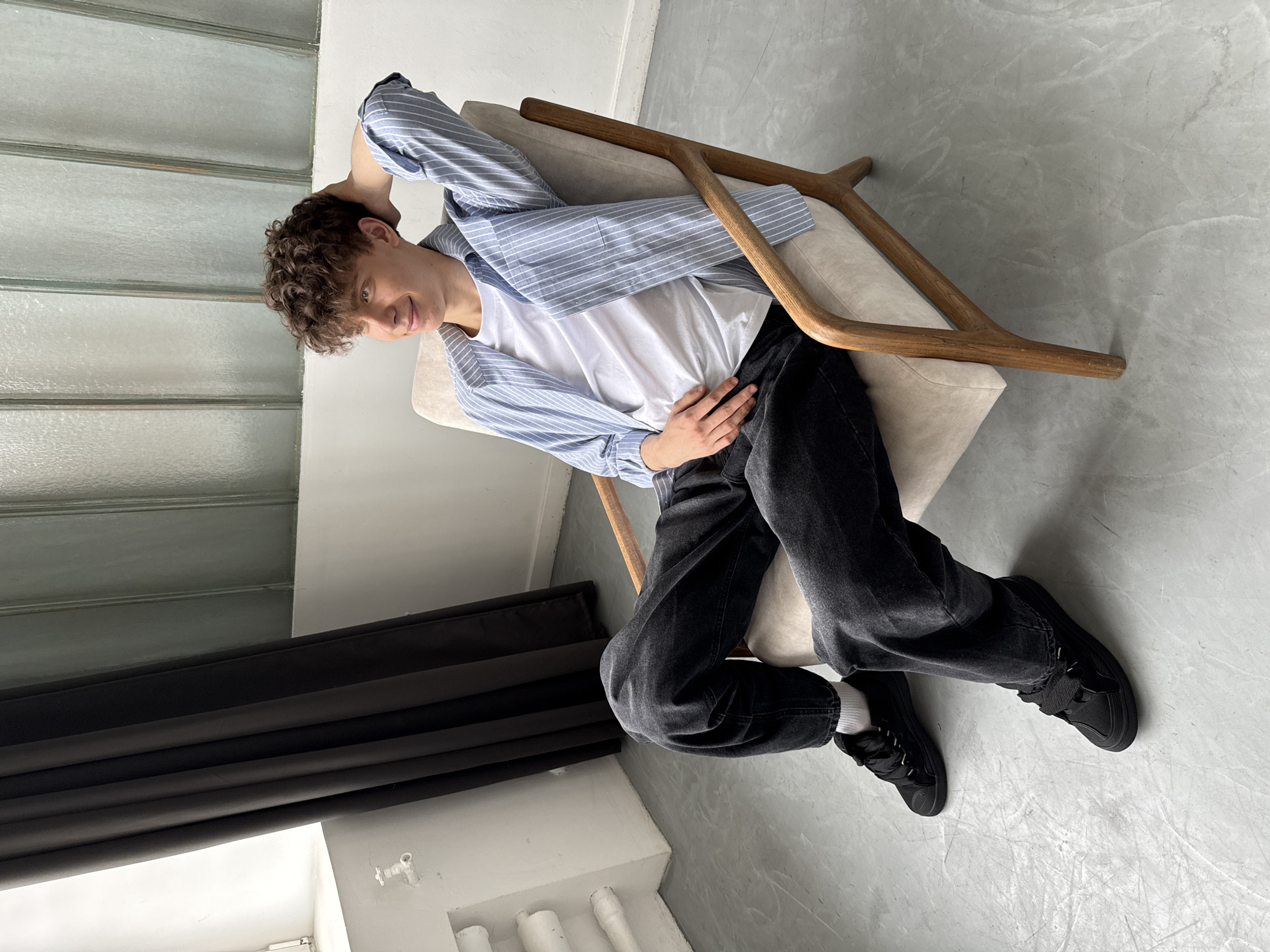 A young man is casually reclined in an arm chair, facing the camera. He has curly brown hair and is wearing a white t-shirt, a light blue and white striped button-down shirt, black jeans, and black sneakers. He is resting one hand behind his head and the other on his stomach.
The arm chair has light beige upholstery and a wooden frame with a distinctive Y-shaped design. The room has a minimalist aesthetic with light gray floors and white walls. To the left, there are black drapes or curtains partially visible, and white cylindrical objects are visible at the base of the wall near a window.
It's impossible to determine if this image was taken in Bor, Russia without additional information or visual cues linking the scene to that specific location.