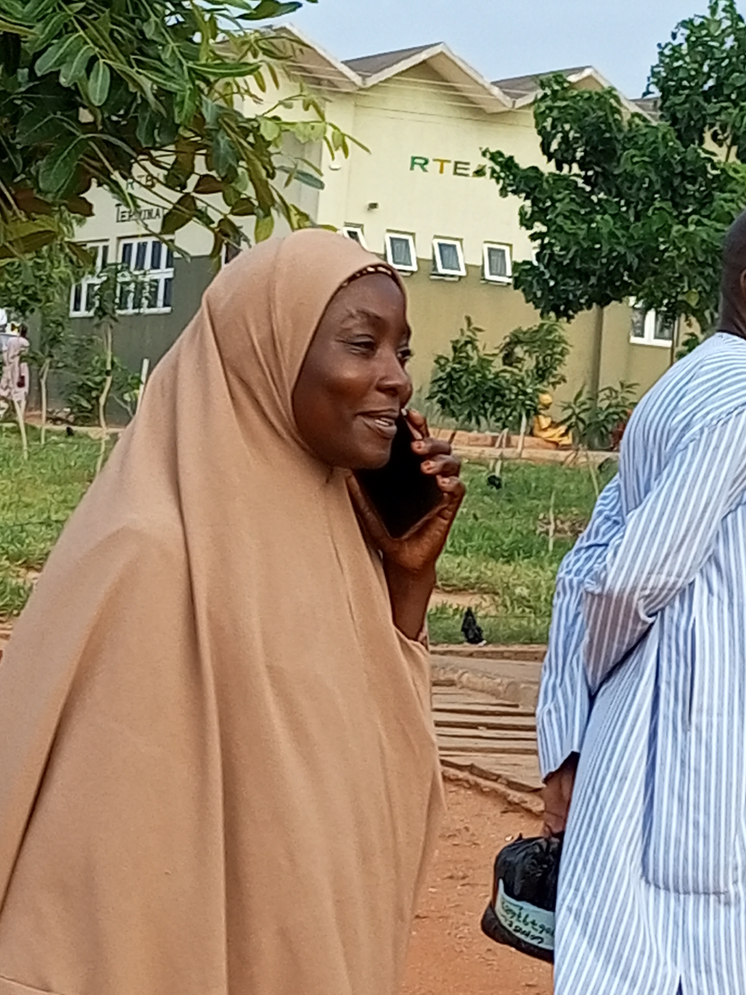 A medium shot shows a woman in a tan hijab on the left, speaking on a dark mobile phone. She has her left hand holding the phone to her ear, and her right hand is visible at her side. To the right of the woman is a partial view of a person wearing a blue and white striped kaftan. In the background, there is a light-colored building with the letters "RTE" visible in green on its facade. There are also trees and green grass in the background. The scene appears to be outdoors, possibly in a public area within the city of Gombe, Nigeria.