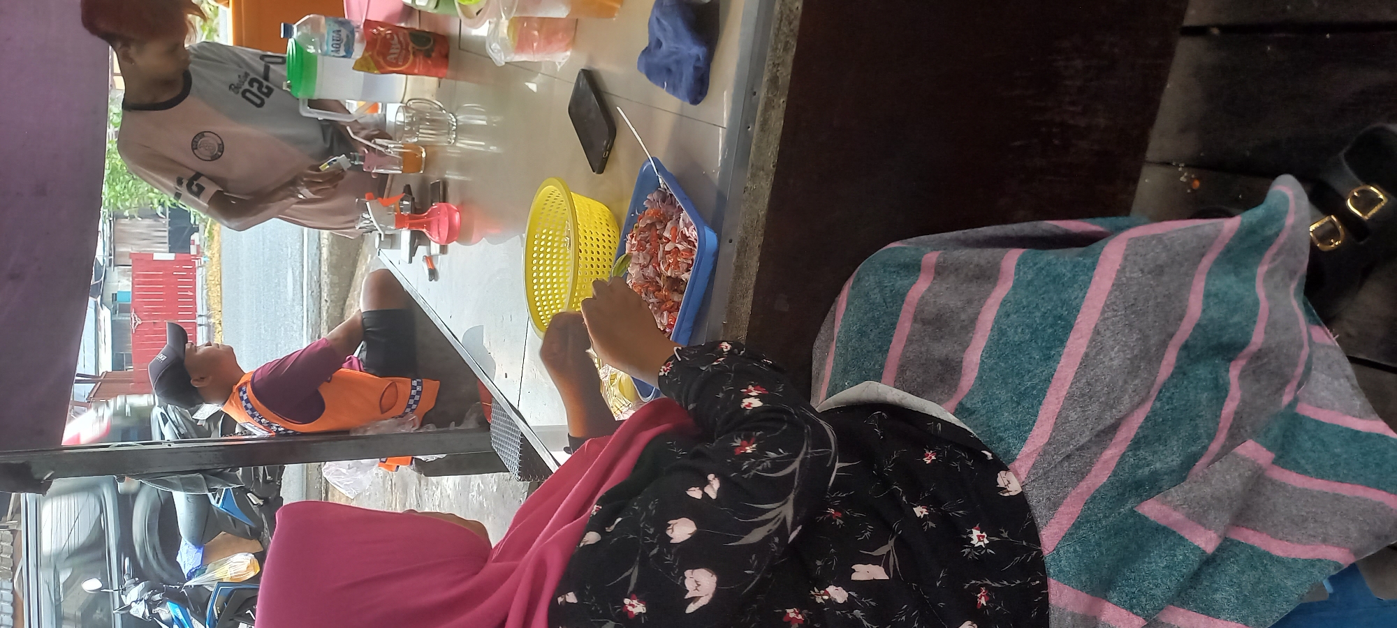 In Melayu, Indonesia, a woman wearing a pink hijab and a floral patterned dress sits at a table, preparing food. In front of her is a blue rectangular tray filled with chopped ingredients, possibly for a dish. Next to the tray is a yellow plastic colander. On the table, other items are visible, including a plastic water bottle, a pitcher, glasses, and a pink spray bottle. To the left of the woman, a man wearing an orange vest and a black baseball cap sits with his back to the viewer. Further to the right, another person is visible from the back, wearing a light-colored t-shirt. The scene appears to be an outdoor food stall or eating area, with a motorcycle parked to the left. The table is placed under a canopy, offering shade.