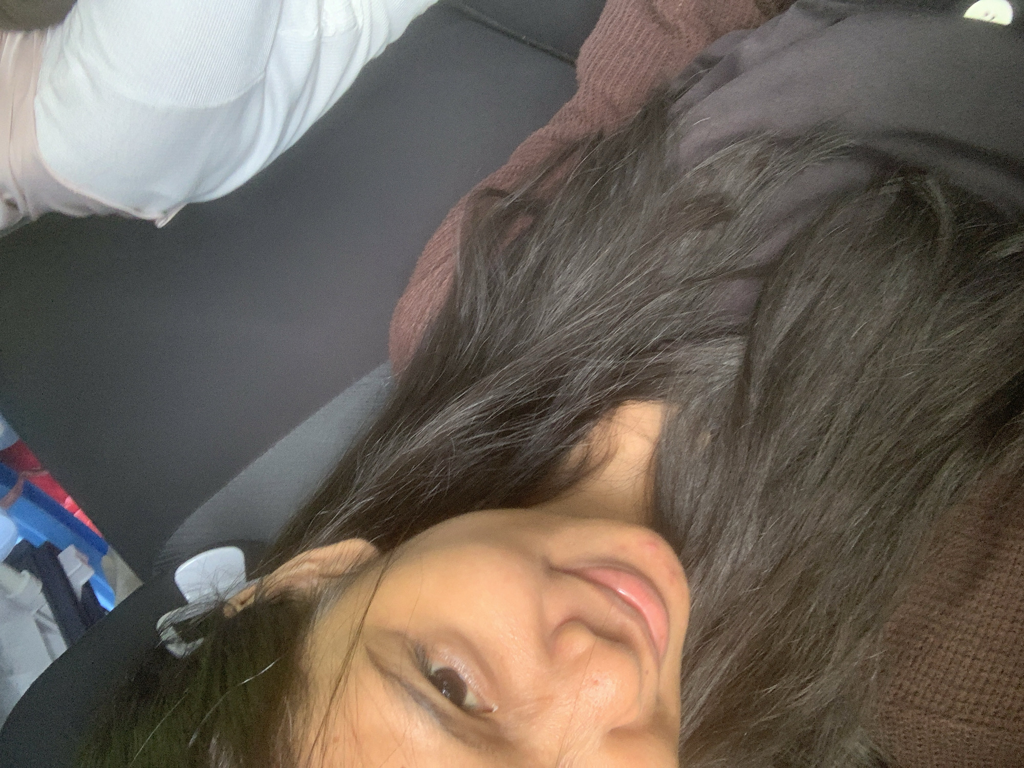 A young woman with dark hair is reclining. She has light brown skin and dark eyes. She is wearing a dark-colored top over a brown sweater or jacket. Some of her hair is spread out around her face. In the background, there is a light-colored garment, a dark grey surface, and a hint of blue and red cloth or materials. The location within the city of Samarinda, Indonesia, cannot be determined from the image.