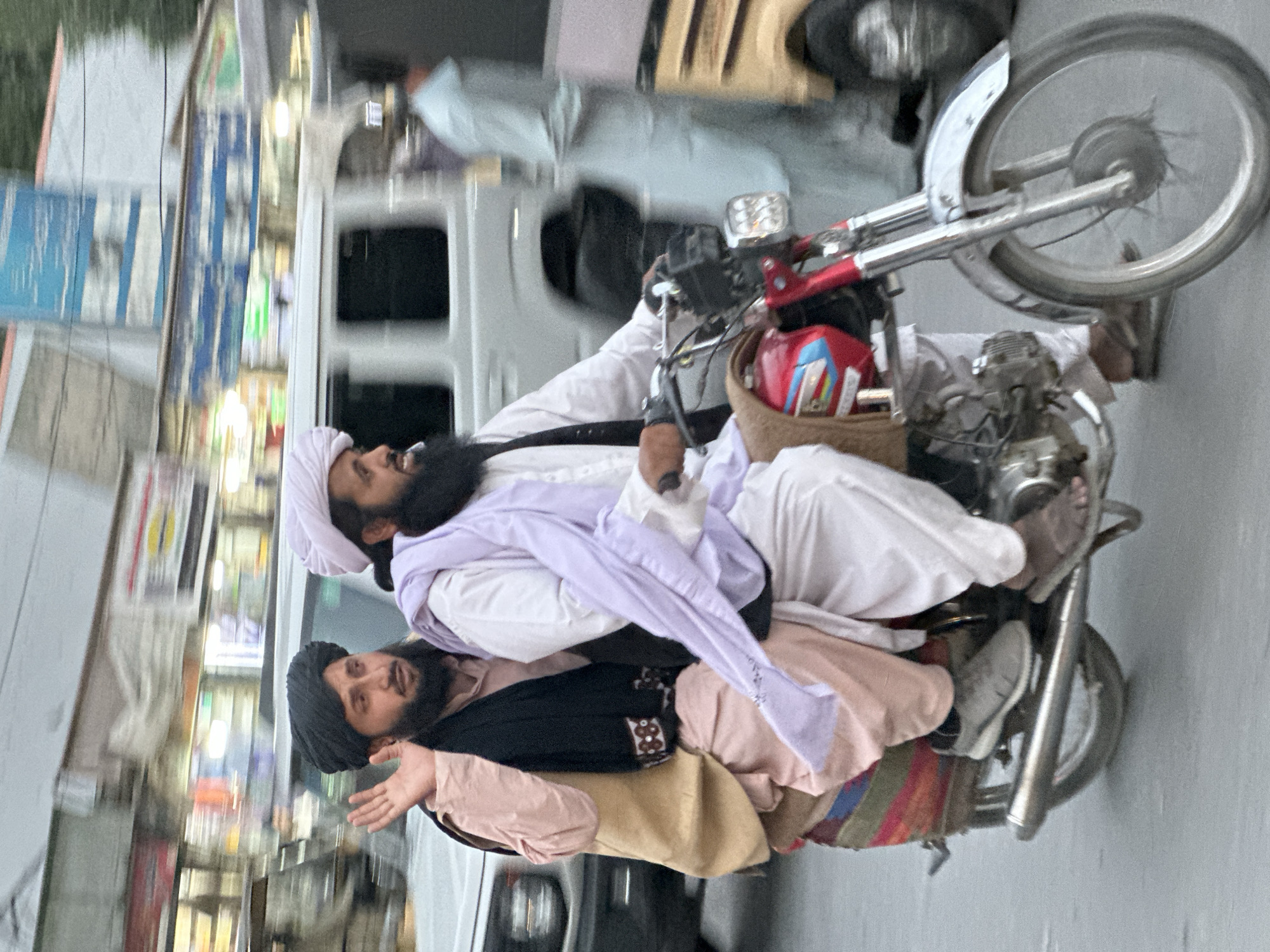 Two men are riding a motorcycle in Quetta City Tehsil, Pakistan. The first man, who is driving, is wearing a white turban, a white shirt, and a lavender scarf. The second man is wearing a black turban, a pink shirt, and a black vest. The motorcycle is old and has a red, blue, and yellow helmet strapped to the back. The background shows blurred storefronts and vehicles, indicating an urban setting.
