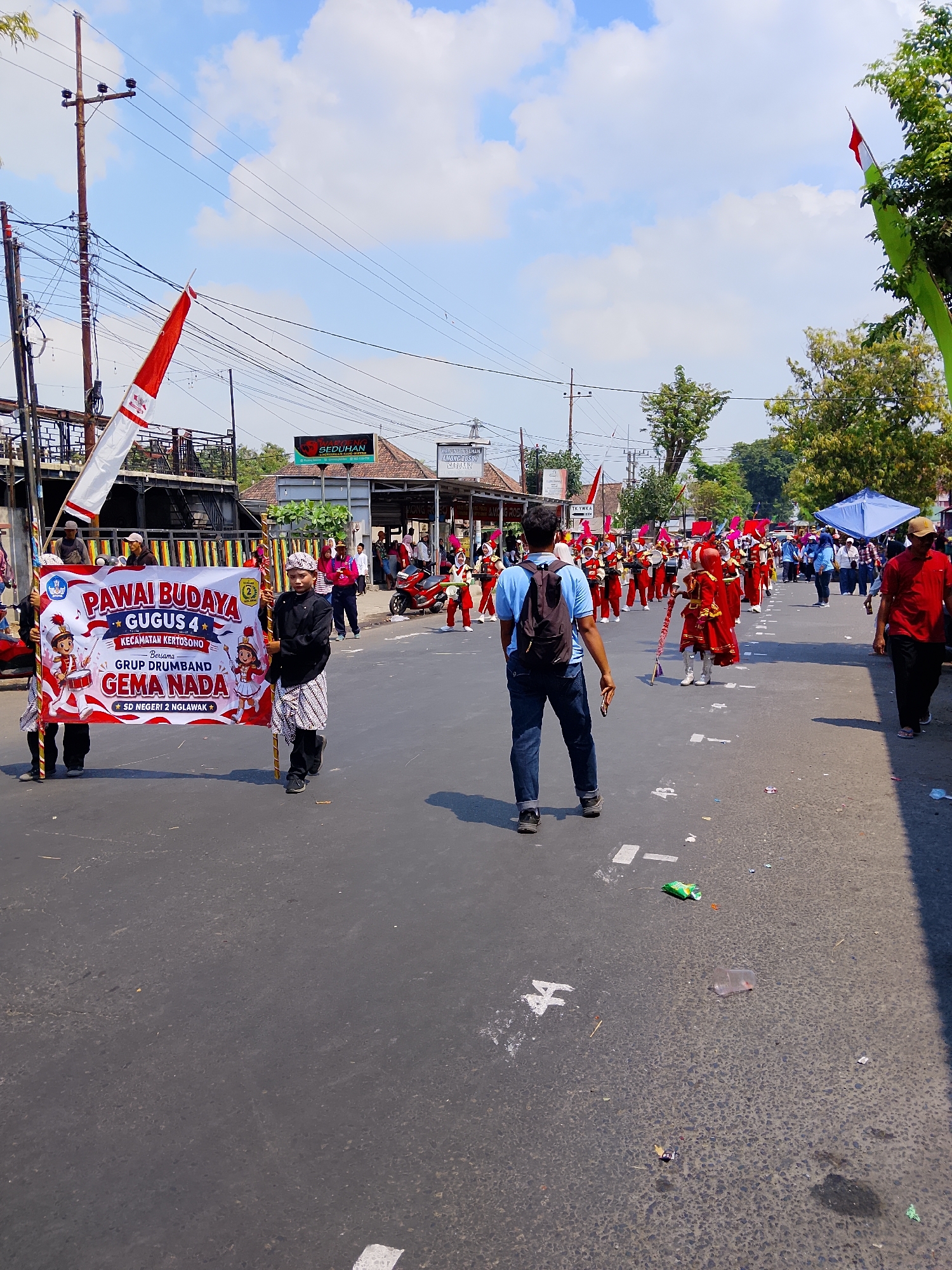 The image depicts a street scene in Kertosono, Indonesia, likely during a cultural parade or event. In the foreground, two individuals are holding a large banner that reads "PAWAI BUDAYA GUGUS 4 KECAMATAN KERTOSONO Bersama GRUP DRUMBAND GEMA NADA SD NEGERI 2 NGLAWAK." This translates to "Cultural Parade, Group 4, Kertosono District, Together with Gema Nada Drum Band Group, State Elementary School 2 Nglawak."
Behind the banner holders, a drum and bugle corps, dressed in red and white uniforms, marches down the street. They are carrying musical instruments such as drums and what appear to be brass instruments. Spectators are visible on both sides of the street and further down the road.
The environment includes a paved road with markings on the surface, overhead power lines, trees lining the street, and buildings in the background, one of which has a sign that reads "SEDHUHAN." The sky is blue with scattered clouds. There is some litter on the road surface. The overall impression is a vibrant community event taking place outdoors on a sunny day.
