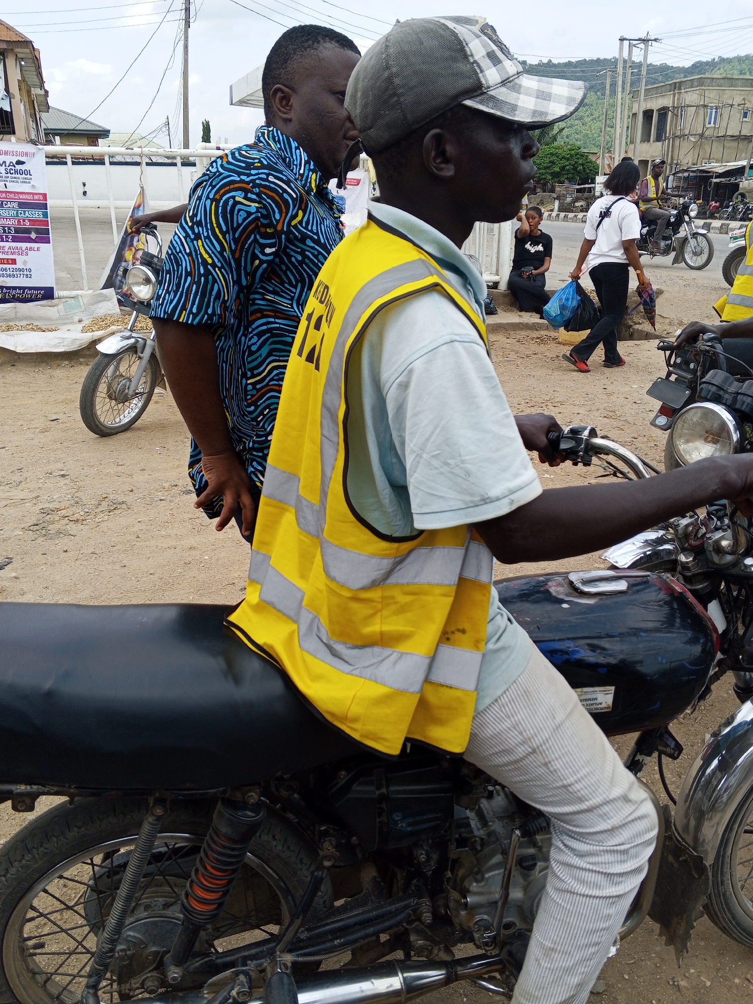 This media file captures a street scene in Zango-ndaji, Nigeria, likely during the daytime, with overcast skies suggesting potential for rain or a generally muted light. The foreground is dominated by a man seated on a black motorcycle, wearing a light blue shirt and a bright yellow safety vest with reflective stripes. He has a cap on his head and appears to be looking off to the right of the frame. His right hand is on the motorcycle's handlebars.
Behind him, another man in a vibrantly patterned blue and black shirt stands next to a white fence, holding a poster or sign. The poster, partially visible, contains text in English, including "ADMISSION!!!", "MA SCHOOL", and mentions of "NURSERY CLASSES" and "PRIMARY". Further back, a woman walks across the street carrying two blue plastic bags and an umbrella. She is wearing a white t-shirt with "AD...high" visible on the back, black pants, and red shoes. Another person sits on a ledge to the left, wearing a black t-shirt and skirt.
In the background, more motorcycles are visible, along with buildings and power lines, indicative of a busy urban or semi-urban environment. The ground is dusty, a common characteristic of Nigerian roads. The overall impression is one of daily life unfolding in a bustling community, with a focus on transportation and possibly local businesses or institutions, as suggested by the poster.