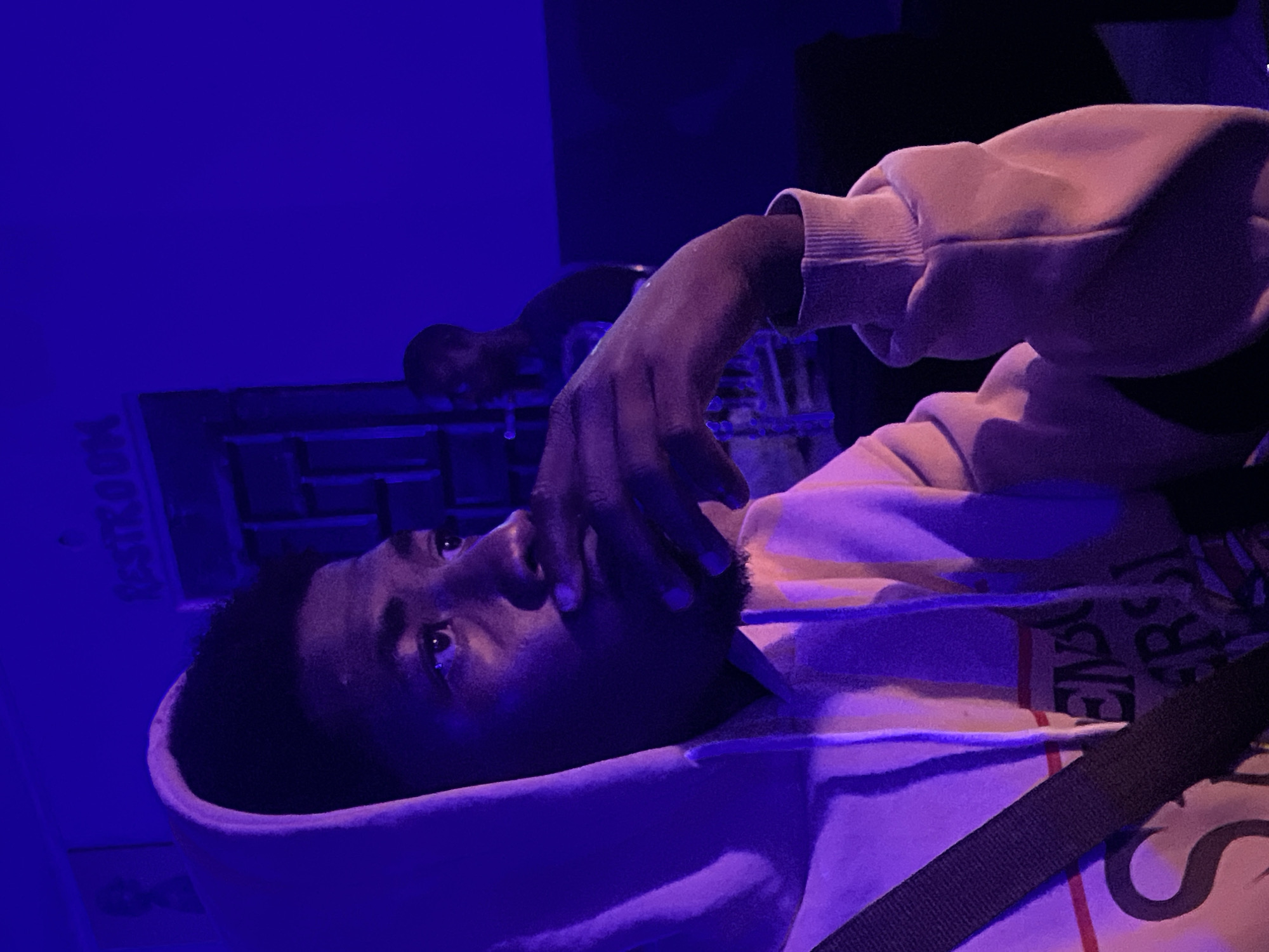 The image shows a young Black man wearing a light-colored hooded sweatshirt, illuminated by purple lighting. He is looking to his right, with his hand on his chin. In the background, another person is partially visible. There is a sign with the word "RESTROOM" visible on the wall. The scene appears to be indoors, possibly in a dimly lit establishment in an urban setting. The overall atmosphere is somewhat moody due to the lighting.