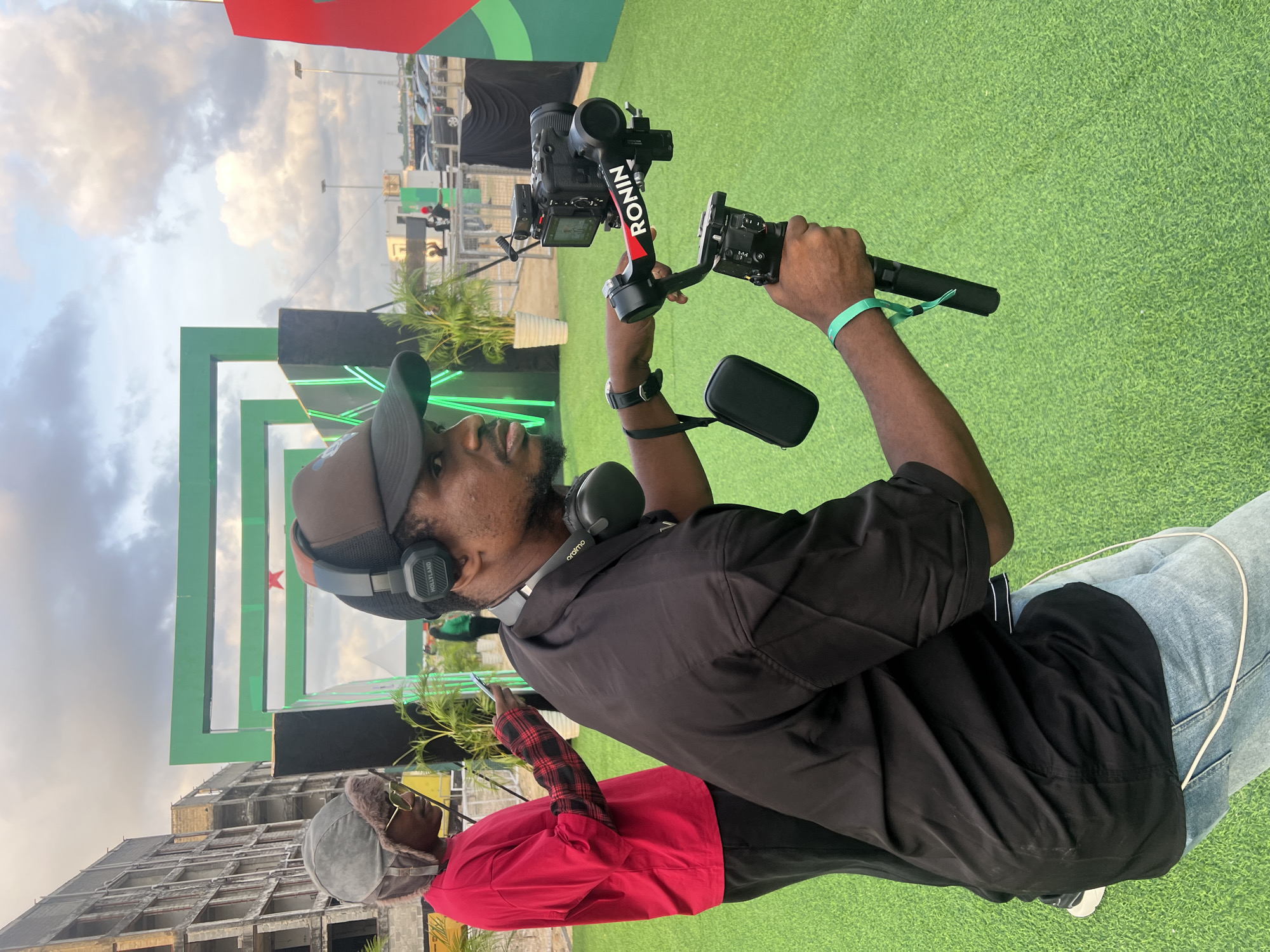 A man in Lagos, Nigeria, operates a Ronin camera stabilizer. He wears a cap, black shirt, jeans, and headphones. Another individual in a red shirt stands partially in the foreground. The backdrop includes an architectural structure, green turf, and a structure with a logo and green lights. A building is visible in the background.