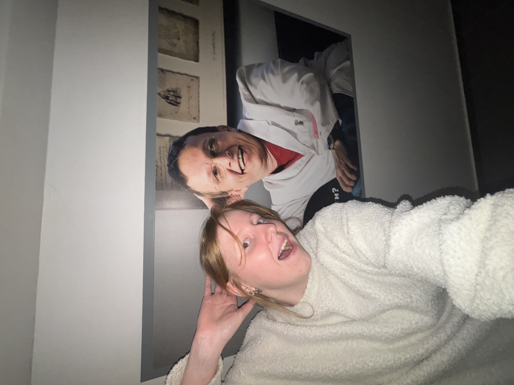 A person wearing a white sweater is in the foreground looking up at the camera and posing. Behind the person is a framed picture mounted on a light-colored wall. The picture depicts a man wearing a white lab coat and smiling. There are also sketches of human anatomy in the background of the picture. Based on the subject of the pictures in the background, the location is likely the Perm Museum of Medical History in Perm, Russia.