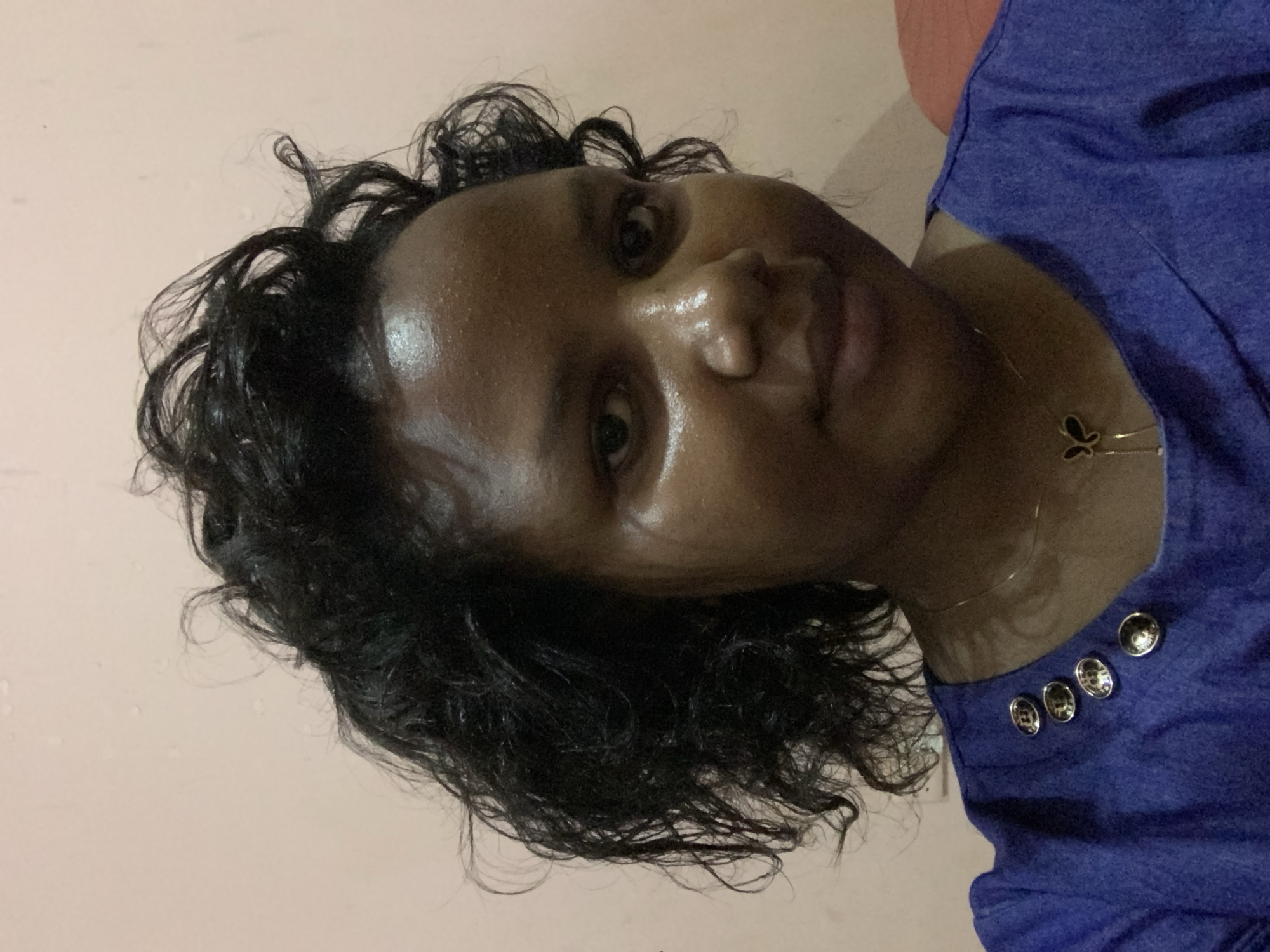 A medium shot shows a person with dark skin, appearing to be female, with wet, curly black hair. Her face is shiny, and she is looking at the camera. She is wearing a royal blue shirt with four round, silver buttons visible on the shoulder. A delicate gold necklace with a butterfly pendant is around her neck. The background is a light peach color. There is no verifiable information in the image to identify it as being in Dar es-Salaam, Tanzania. The picture's origin and location are not discernible from the provided visual elements.