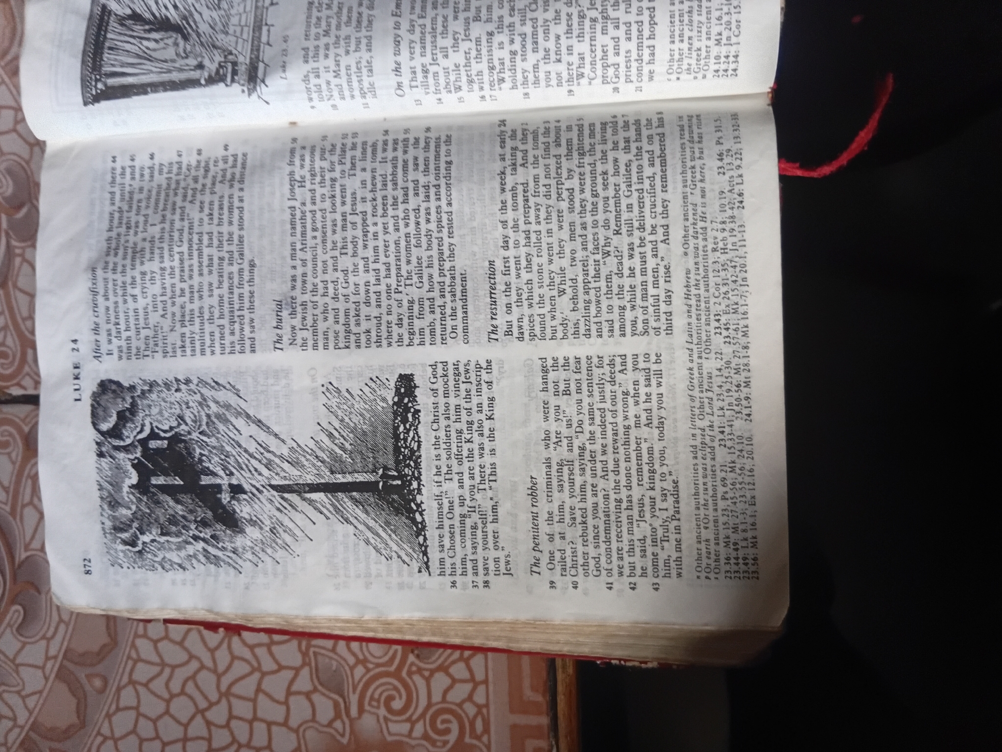 This media file displays an open book, specifically a Bible, with visible text and illustrations. The page is predominantly white, with black text printed in two columns. The left column has a large illustration at the bottom depicting a cross with dark, dramatic clouds and rain-like lines. The text appears to be from the Book of Luke, as indicated by the chapter and verse numbers visible.
The illustration of the cross suggests a scene related to the crucifixion or a significant religious event. The surrounding text discusses the crucifixion, the burial of Jesus, and his resurrection, referencing events like the darkness over the land and the preparation of spices. There are also footnotes at the bottom of the page containing biblical references and notes on ancient authorities.
The book itself has a red cover on the right side, peeking into the frame. The lighting suggests an indoor setting, possibly during the day, with a shadow obscuring the right side of the image. The angle of the photo indicates it was taken from above, looking down at the open Bible. The overall mood conveyed by the illustration and the subject matter of the text is somber and significant, befitting the themes of death and resurrection.