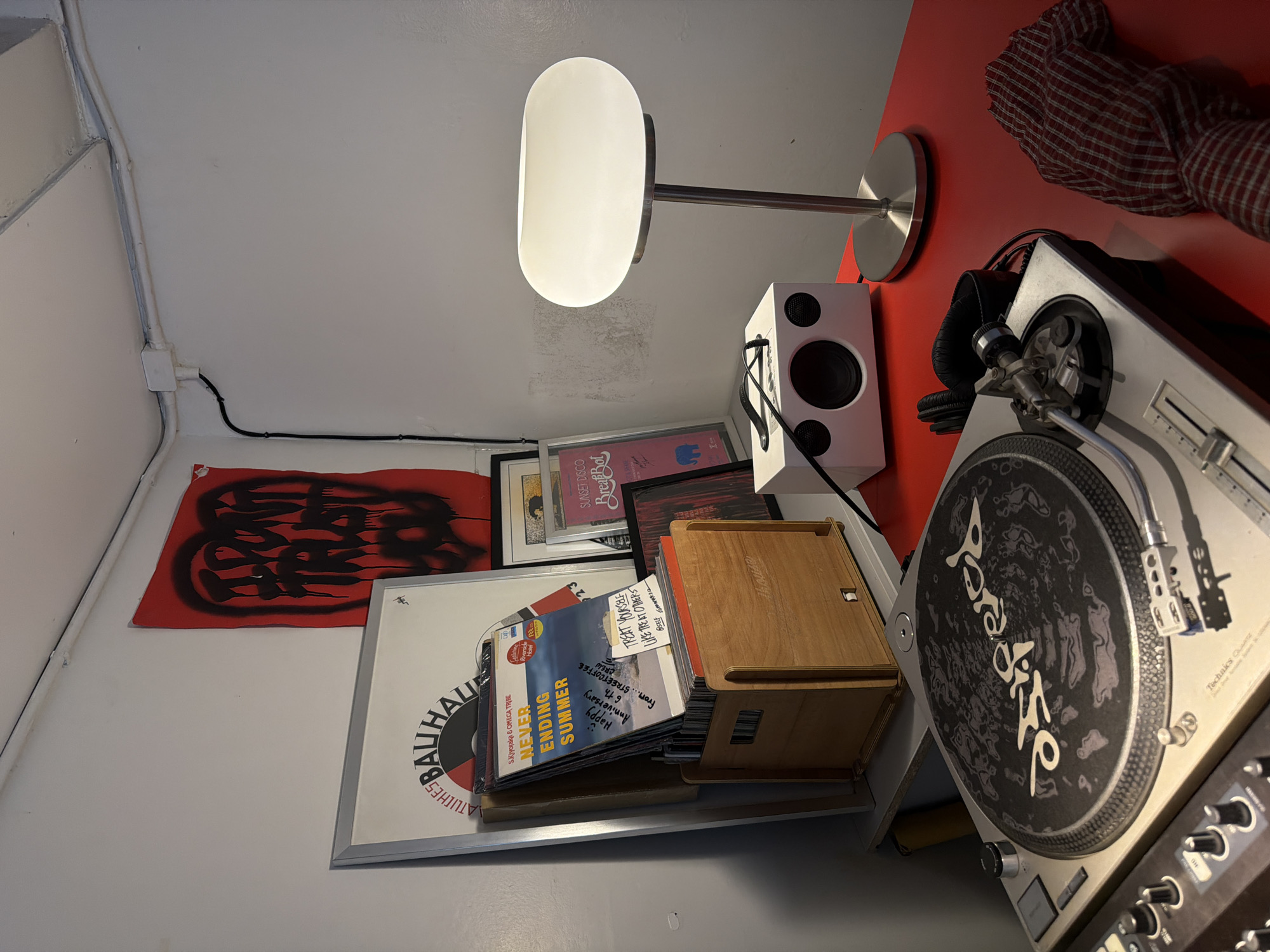 This media file captures a close-up, low-angle view of a collection of items arranged on a red surface and a white shelf against a white wall. The scene appears to be an indoor setting, possibly a home studio or a music enthusiast's space, given the presence of audio equipment and records.
The main objects include a modern white speaker, a pair of black headphones, a turntable with a record on it, and a small wooden storage box filled with records or CDs. A large, minimalist white lamp with a metal stand is positioned prominently in the upper center of the frame, casting a soft light. To the left, a red cloth with a subtle checkered pattern is draped over the red surface. On the wall, a red banner with black abstract graffiti-like lettering is displayed, along with framed artwork or posters. A stack of items, likely magazines or album covers, leans against the wooden box, with some visible text including "NEVER ENDING SUMMER" and "Happy Anniversary 6th from... STREETCOFFEE CREW." The turntable itself has "PARADISE" printed in a stylized font on the record mat. Another visible text on the turntable is "Technics". The overall mood is one of organized clutter, suggesting a passion for music and art. The lighting is somewhat dim, suggesting it could be evening or an indoor environment with controlled lighting.