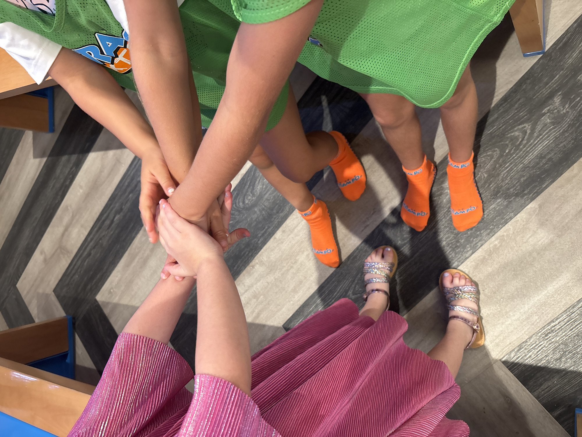 A high-angle photograph captures four individuals, identified as children, engaged in an activity within an indoor facility in Dubai, United Arab Emirates, consistent with an indoor play area or activity center. The subjects' arms extend towards the center of the frame, where their hands are stacked, suggesting a collective huddle or game initiation. On the left side, two individuals are dressed in a reflective fuchsia-pink pleated ensemble. One individual's feet are visible, adorned with metallic multi-strap sandals and light-colored nail polish. On the right, two individuals wear green mesh sports jerseys over white t-shirts. Both display bright orange grip socks, one pair clearly showing 'CAMPO' lettering on the soles, covering their feet and lower legs. The flooring is a contemporary chevron pattern, featuring alternating light and dark grey wood-effect planks. A wooden structure with blue detailing, possibly part of a play equipment or furniture, is partially visible in the upper background.