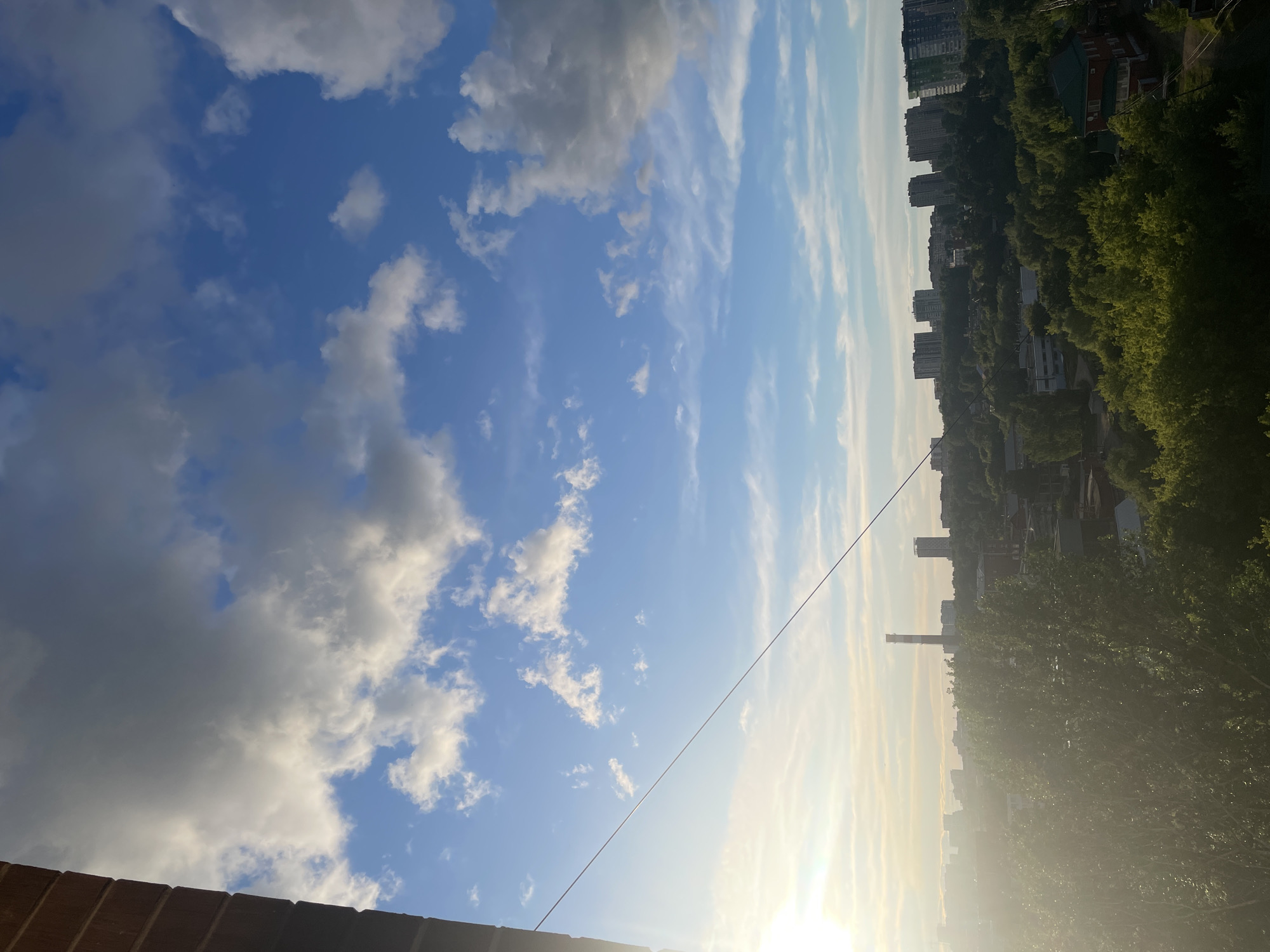 The image depicts a sky filled with scattered cumulus clouds over Izhevsk, Russia. Below the sky, the cityscape includes buildings and dense green trees. A tall smokestack is visible. A power line stretches across the sky. The photo is taken from a high vantage point, such as a balcony or upper-story window, with a brick ledge visible in the foreground. The sun shines brightly, creating a slight haze in the distance.
