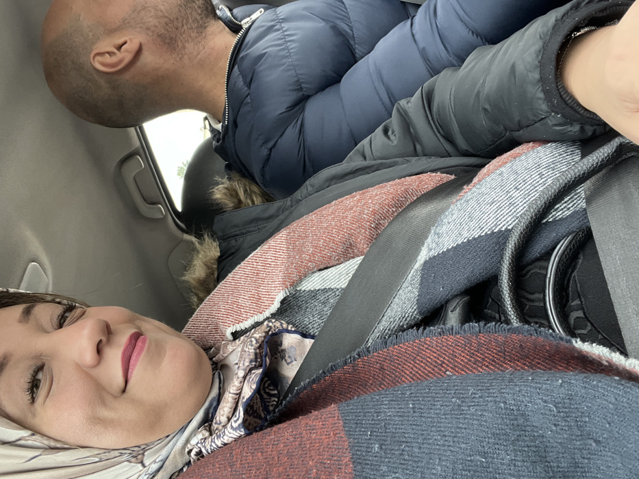 Inside a vehicle, a woman with light skin, wearing a patterned headscarf and pink lipstick, smiles into the camera. She is surrounded by blankets and wearing a seatbelt. Partially visible above her is a man with a shaved head, wearing a blue puffer jacket. The car interior includes a grey ceiling, a window, and a handle.