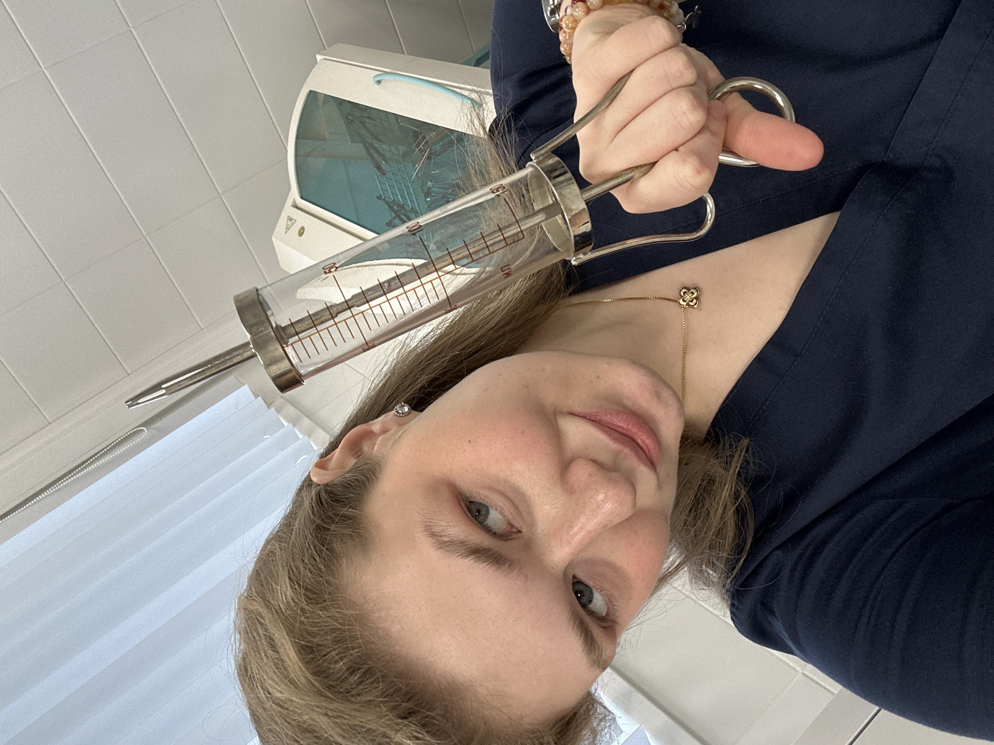 The image shows a woman holding a large syringe. The syringe is glass with metal components, marked with volume measurements. The woman is wearing a dark blue top and a gold necklace with a clover-shaped pendant. Her hair is light brown, and she has light skin and blue eyes. The background includes a white tiled wall and a medical device with a blue-tinted screen. The location is inferred to be in Kazan, Russia.