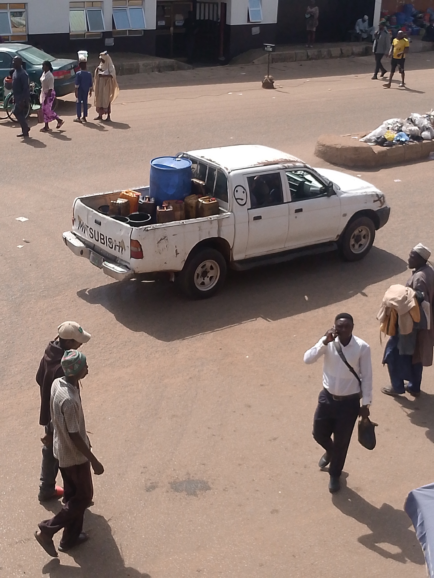 This image captures a street scene in Bukuru, Nigeria, under what appears to be bright daylight, suggested by the strong shadows. The central focus is a white Mitsubishi pickup truck loaded with various containers, including a large blue barrel and several smaller yellow and brown containers. The truck also has a crudely drawn smiley face on its rear passenger side door.
In the foreground, three men are visible. Two younger men, one wearing a light-colored plaid shirt and a beanie, and another in a darker jacket and a cap, are walking and appear to be conversing. A third man, dressed in a white shirt and dark trousers, is walking and talking on a mobile phone, carrying a dark bag over his shoulder. To the right of the pickup truck, an older man in traditional attire, carrying a bundle of clothing, is also present.
In the background, several other individuals can be seen, along with a green sedan and a bicycle. The setting appears to be an urban or semi-urban environment, with buildings lining the street. The overall atmosphere is that of a busy day with people engaged in various activities.