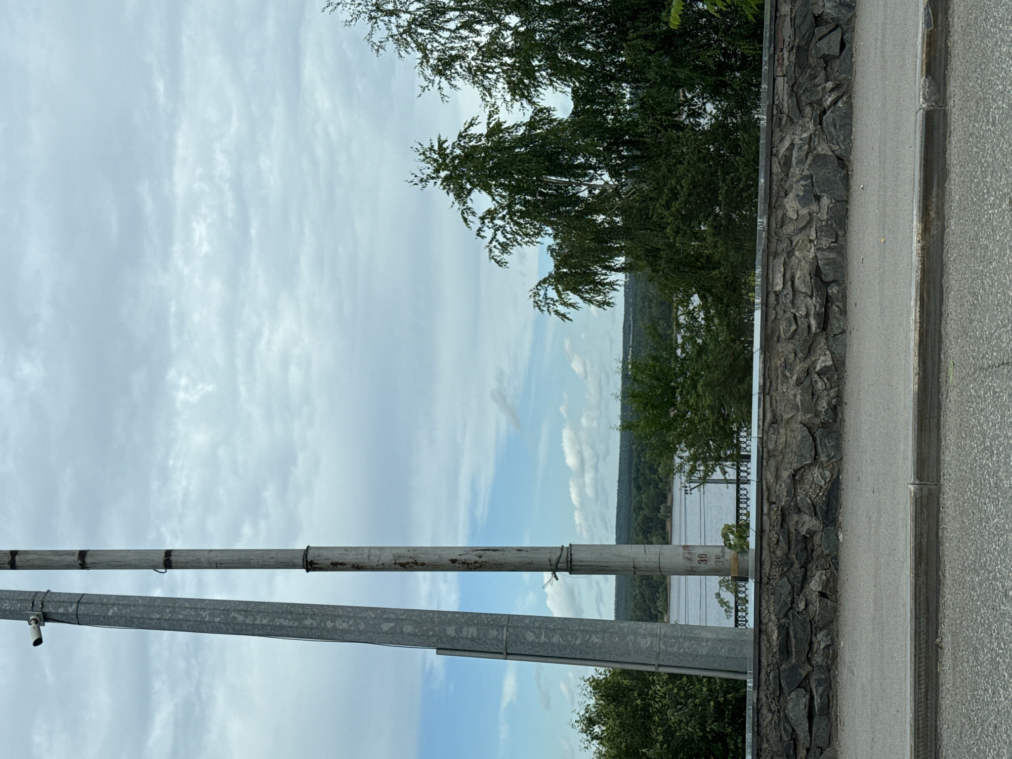 The image shows a cloudy sky over a body of water, likely the Kama River in Perm, Russia. In the foreground, there is a stone wall topped with a metal railing, and a paved area, possibly a sidewalk or road. Two tall poles stand vertically on the left side of the frame; the left pole is made of galvanized metal and has a surveillance camera attached near the top. The pole to its right is older, made of concrete, and shows signs of rust and wear, with some markings that appear to be numerical (possibly "30" and "39"). Behind the wall, dense green trees line the riverbank, with a distant, hazy treeline on the opposite shore. The overall scene is overcast and appears to be a public space overlooking the river.