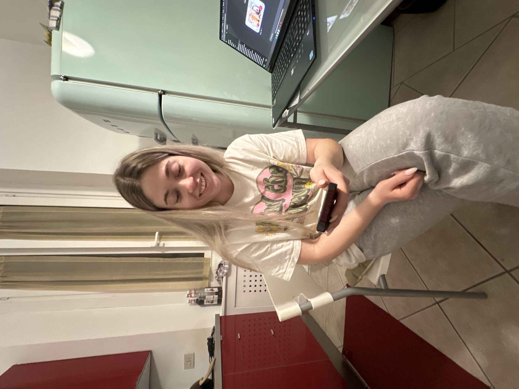 A young woman with long blonde hair is depicted smiling brightly while seated indoors, likely in a kitchen or a combined living/kitchen area in Milan, Italy. She is wearing a light-colored t-shirt with a floral graphic design and light grey sweatpants, suggesting a casual and relaxed atmosphere. She is holding a dark smartphone in her left hand, her gaze directed towards it or just beyond it, indicating she is engaged with its content.
To her left, a distinctive light green, retro-style refrigerator (reminiscent of Smeg appliances) stands prominently. Adjacent to the refrigerator, a black laptop is open on a glass surface, displaying a screen with what appears to be a graphical interface or an image. The background on the right features a window with horizontal blinds and a red cabinet or countertop below it. The floor is tiled with light-colored square tiles.
The indoor lighting suggests it is likely evening or nighttime. The woman's joyful expression implies a moment of leisure and happiness, possibly while browsing content on her phone after or during a period of work/study on the laptop. No clear, readable text is discernible on her clothing or the visible objects, apart from potential interface elements on the laptop screen that are too small to read.